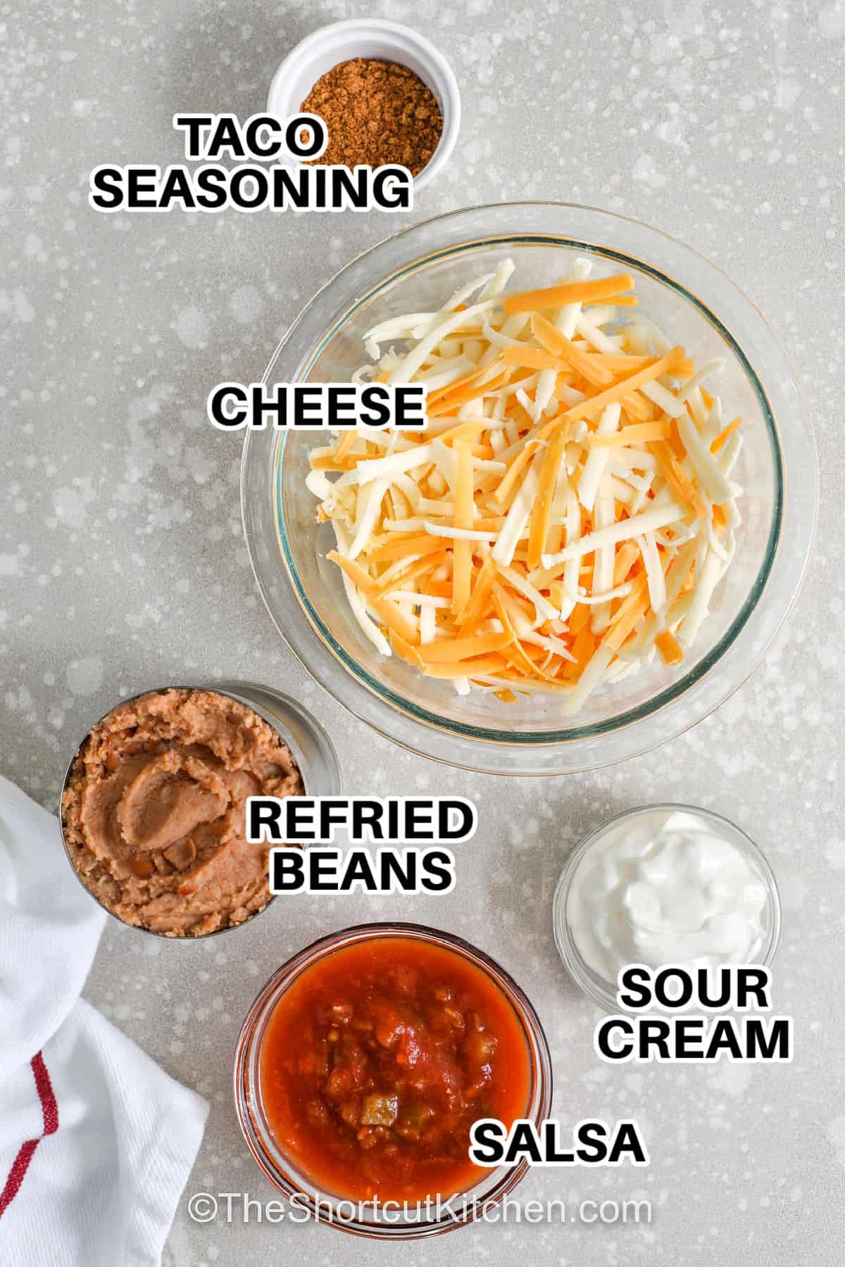 Refried bean dip is a warm party appetizer that is easy to prepare. Refried beans are mixed with salsa, taco seasoning, and sour cream before baking under a layer of melted cheese. This easy hot dip recipe is perfect with tortilla chips and makes a great Tex-Mex snack for game day or potluck gatherings. Keep this baked bean dip in your recipe collection for simple entertaining and casual meals with family and friends. #theshortcutkitchen taco seasoning , cheese , refried beans , salsa and sour cream with labels to make Refried Bean Dip