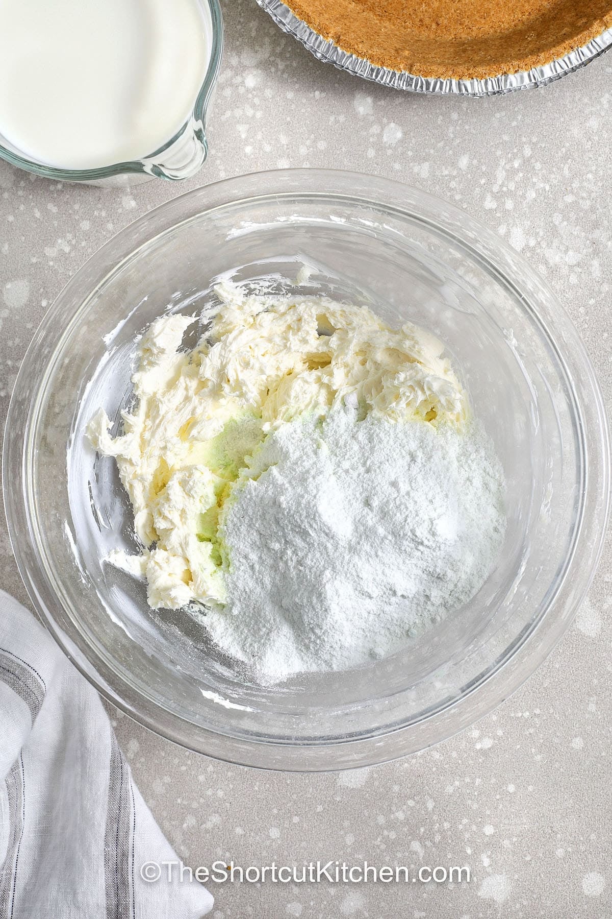 Pistachio pie is the ultimate make-ahead dessert for busy days. This pistachio pudding pie uses instant pistachio pudding mix, cold milk, cream cheese, and whipped topping for a smooth, sliceable no bake pistachio pie. A premade graham cracker crust makes it even faster. Chill overnight for the cleanest cuts, then finish with pistachios or maraschino cherries. #theshortcutkitchen adding pudding mix to bowl to make Pistachio Pie