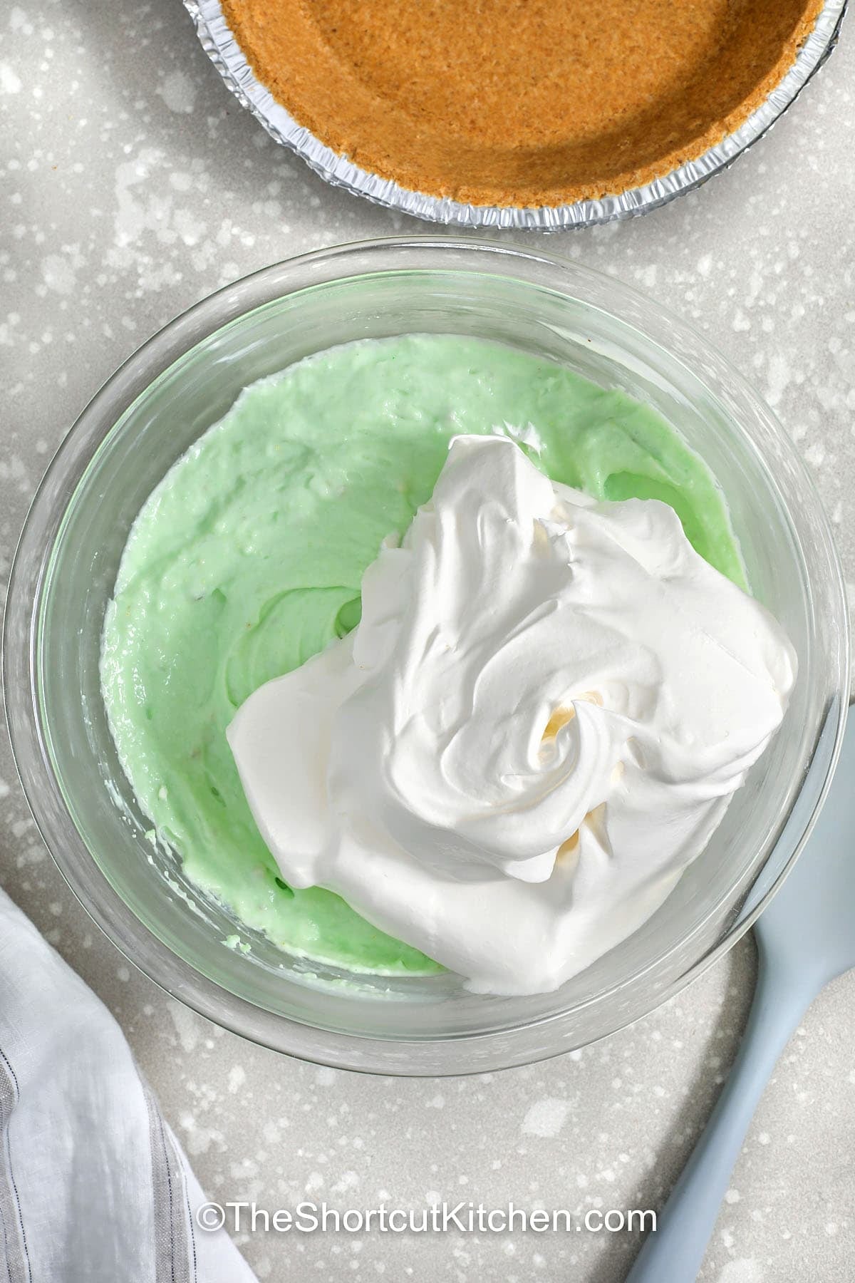 This Pistachio pie is a dreamy no bake pistachio pie that comes together in minutes. Beat cream cheese, mix in instant pistachio pudding, add cold milk, then fold in Cool Whip for a fluffy pistachio pudding pie filling. Spoon into a graham cracker crust and chill until set. Top with crushed pistachios for crunch or cherries for a retro look. #theshortcutkitchen adding whipped topping to bowl to make Pistachio Pie