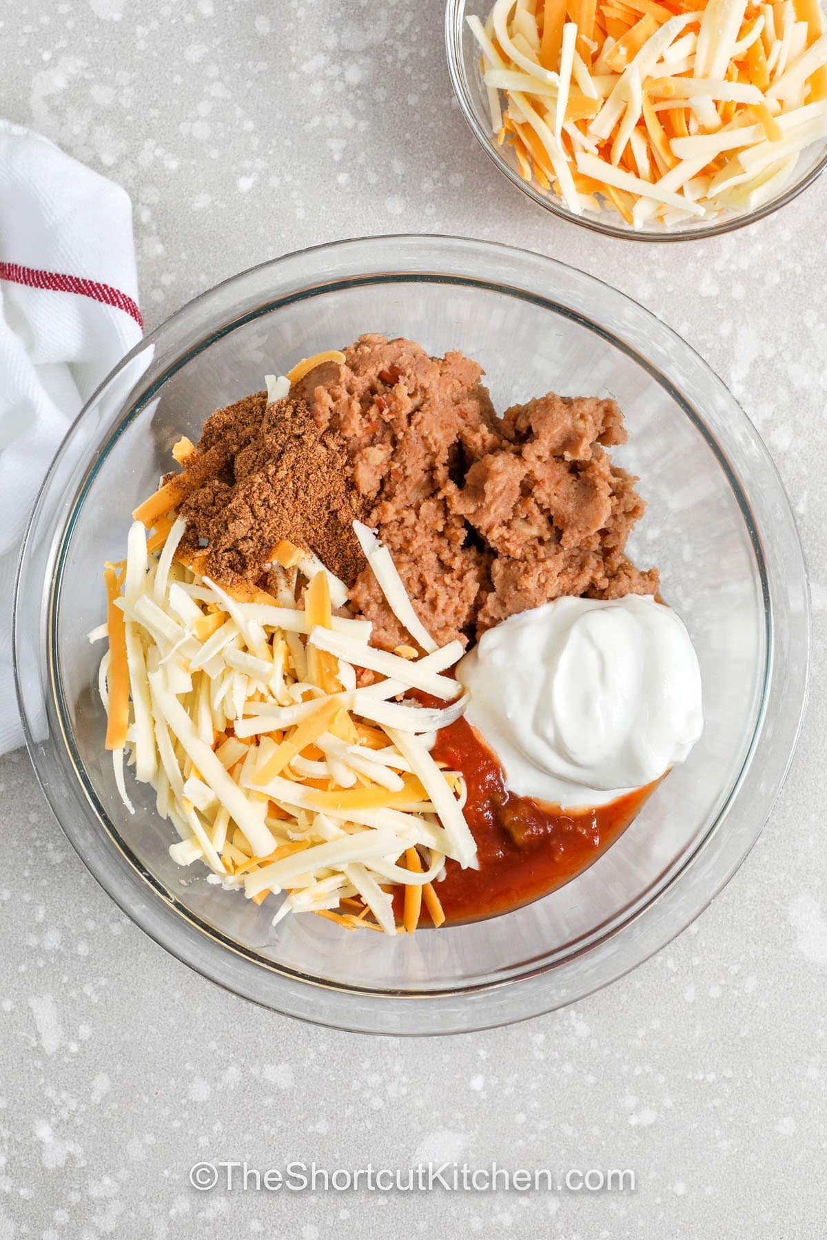 Refried bean dip is a simple baked appetizer that is full of Tex-Mex flavor. Refried beans blend with salsa, sour cream, taco seasoning, and melted cheese to create a warm party dip that is easy to share. This hot bean dip is perfect for tortilla chips and works well for potluck recipes or game day snacks. Keep this easy appetizer recipe ready for gatherings when you want a comforting dip that everyone will enjoy at the table. #theshortcutkitchen mixing refried beans with cheese , salsa , sour cream and seasoning