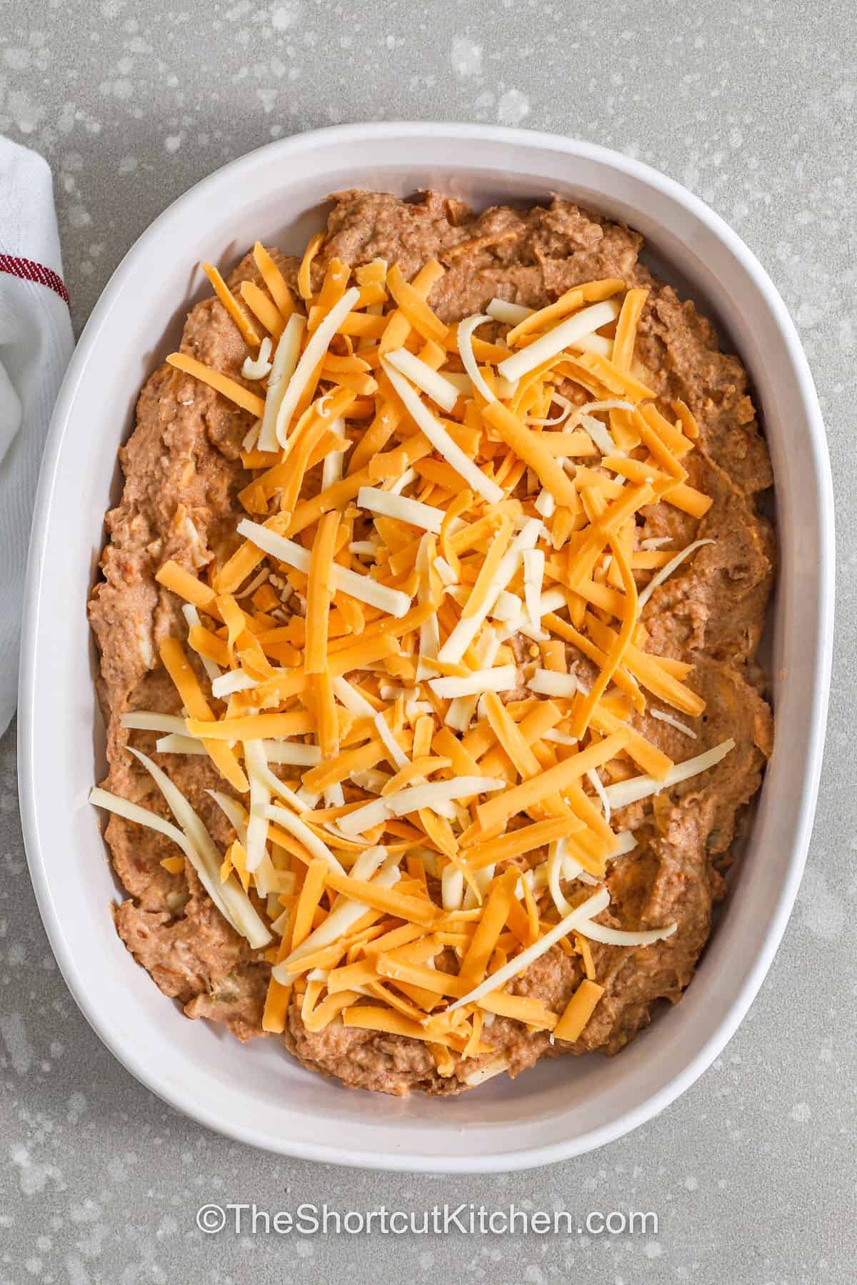 Refried bean dip makes a warm and cheesy Tex-Mex appetizer for sharing. Refried beans are mixed with salsa, sour cream, and taco seasoning, then baked under a layer of melted cheese. This easy bean dip is perfect with tortilla chips or crackers and works well for game day snacks or potluck dishes. Serve this baked dip when you need a simple party appetizer that brings bold flavor and plenty of scoops for friends and family to enjoy together. #theshortcutkitchen adding cheese to Refried Bean Dip