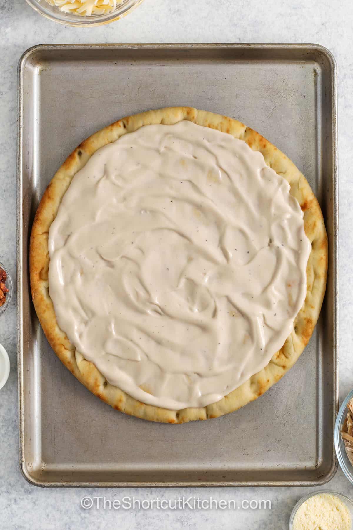 Alfredo pizza is a simple way to change up pizza night with creamy Alfredo sauce instead of tomato sauce. A pre-baked crust holds layers of mozzarella, rotisserie chicken, and bacon bits for a hearty slice. Parmesan cheese and Italian seasoning add extra flavor on top. This easy chicken Alfredo pizza recipe bakes up golden and bubbly and is a great homemade pizza idea for dinner. #theshortcutkitchen adding alfredo to pizza crust to make Chicken Alfredo Pizza