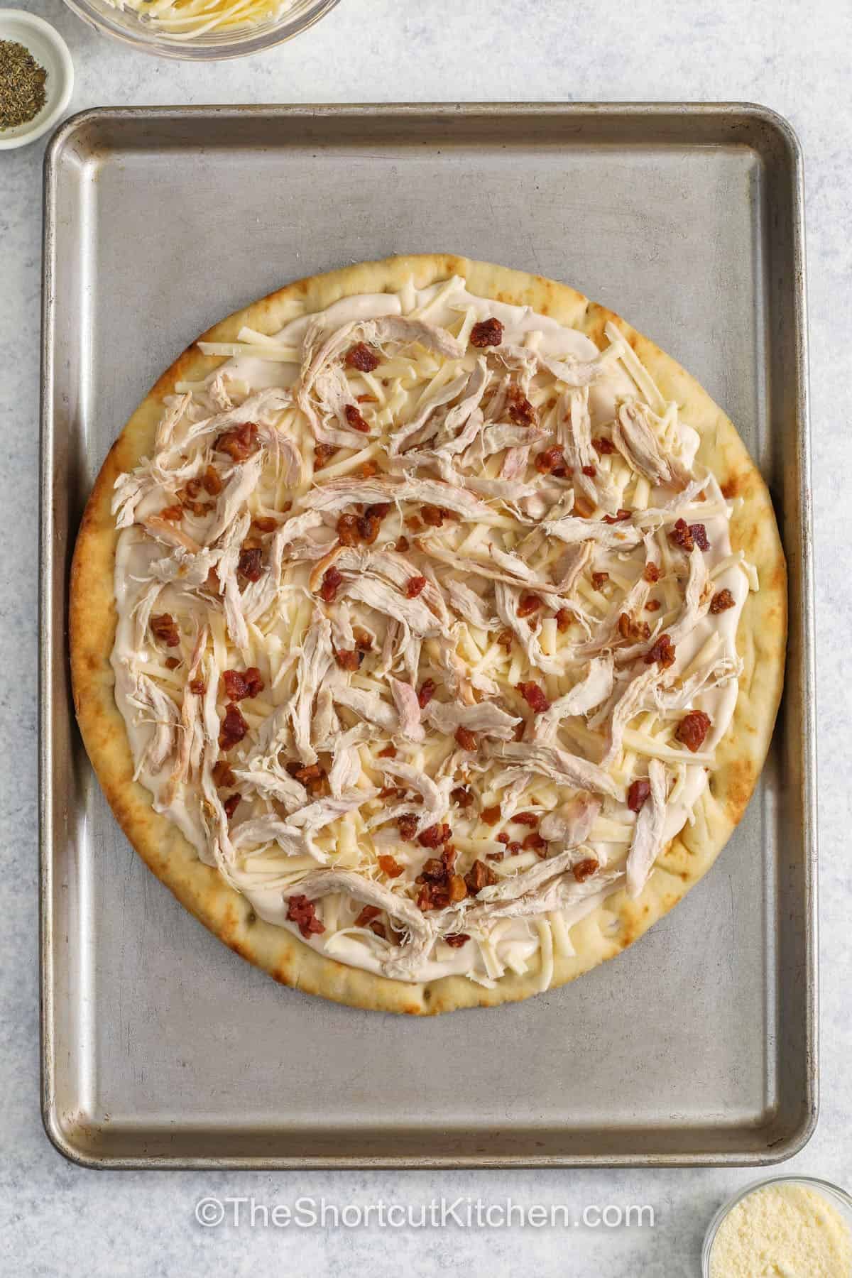 Alfredo pizza brings creamy sauce and cheesy flavor together on a crisp crust for an easy dinner idea. This chicken Alfredo pizza is topped with rotisserie chicken, bacon bits, and a generous layer of mozzarella cheese. A sprinkle of Parmesan and Italian seasoning adds a simple finish that tastes great in every bite. It is a family-friendly homemade pizza recipe that is perfect for weeknight meals. #theshortcutkitchen adding chicken and bacon to pizza to make Chicken Alfredo Pizza