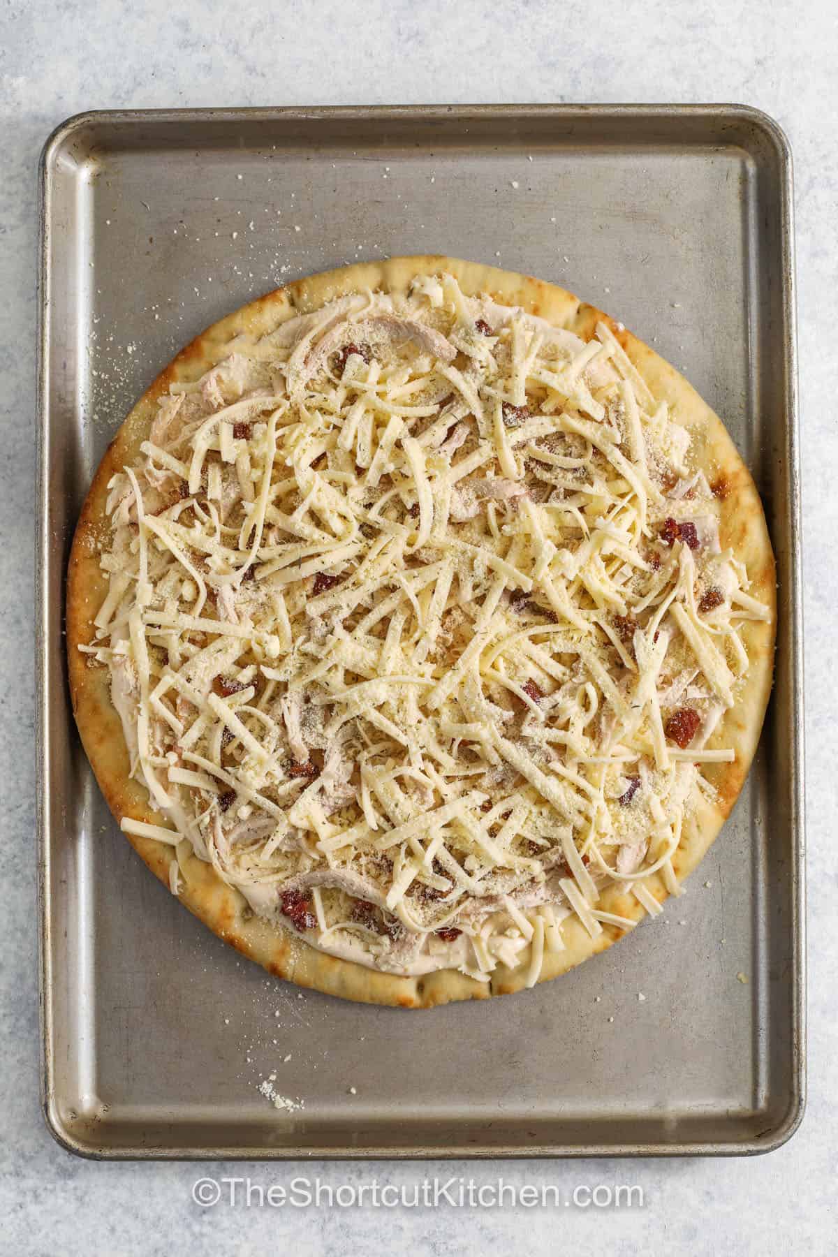 Alfredo pizza is a creamy, cheesy twist on pizza night that everyone enjoys. A crisp pre-baked crust is layered with rich Alfredo sauce, juicy rotisserie chicken, smoky bacon bits, and plenty of mozzarella. A sprinkle of Parmesan and Italian seasoning adds classic flavor to every slice. This easy chicken Alfredo pizza recipe is great for busy nights when you want something warm and homemade from the oven. #theshortcutkitchen adding cheese and seasoning to pizza to make Chicken Alfredo Pizza