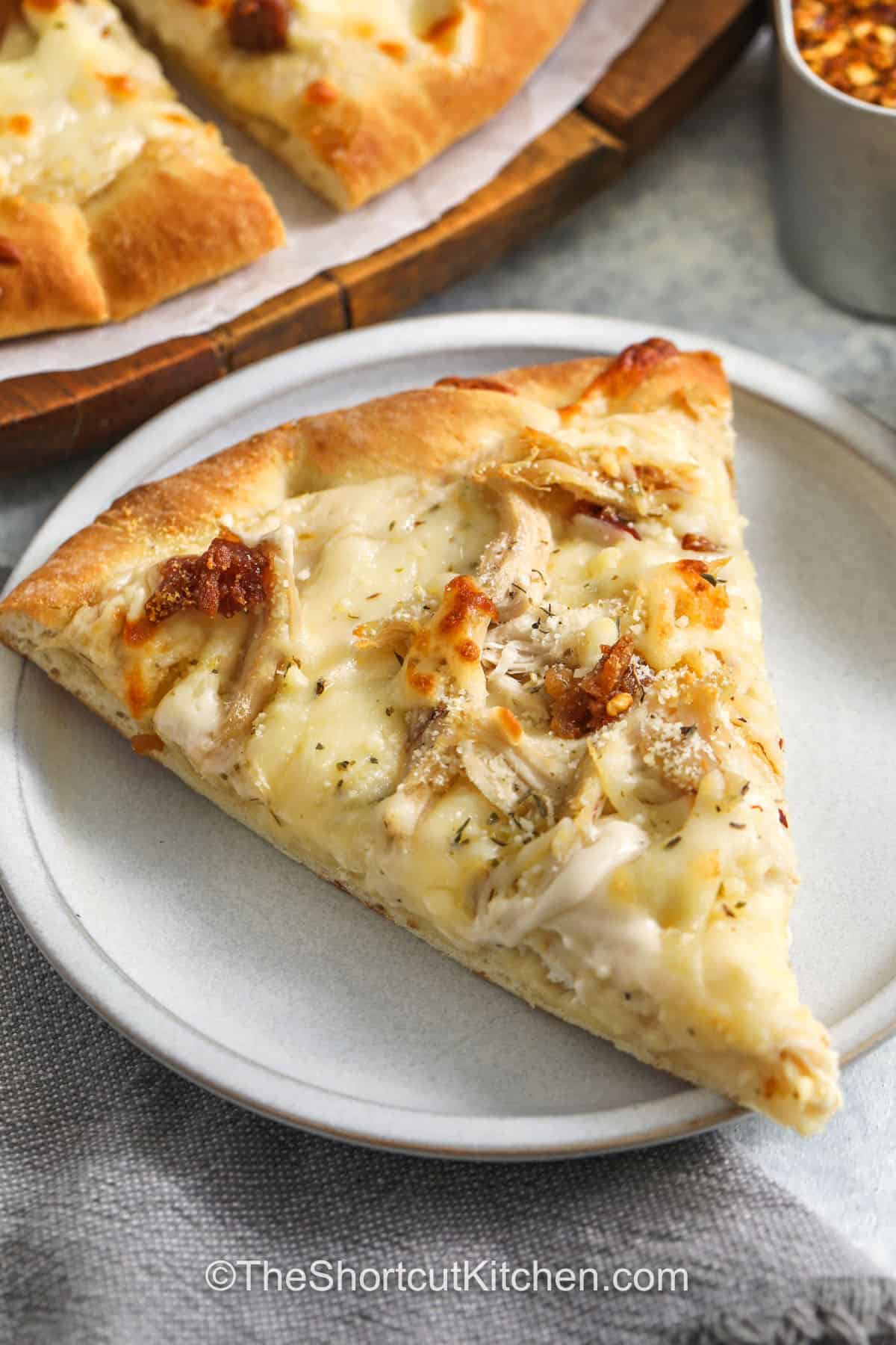 Alfredo pizza turns simple ingredients into a delicious homemade pizza everyone will want again. Creamy Alfredo sauce spreads over a pre-baked crust and is topped with mozzarella, rotisserie chicken, and bacon bits. Parmesan cheese and Italian seasoning add extra flavor to every slice. This easy chicken Alfredo pizza recipe is perfect for a cozy dinner with very little effort. #theshortcutkitchen slice of Chicken Alfredo Pizza on a plate