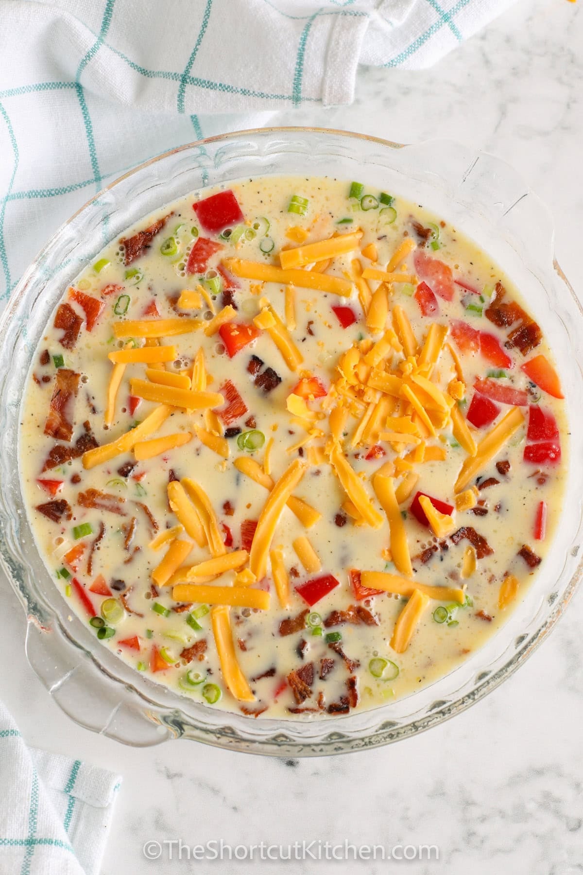 Crustless bacon quiche is an easy savory breakfast recipe made with crispy bacon, cheddar cheese, eggs, milk, Bisquick, green onions, and red pepper. This crustless quiche recipe bakes in one pie plate and comes out fluffy, cheesy, and perfect for breakfast, brunch, or dinner. It is a simple bacon quiche recipe for busy mornings, meal prep, or a holiday brunch spread. #theshortcutkitchen uncooked quiche in a pie plate