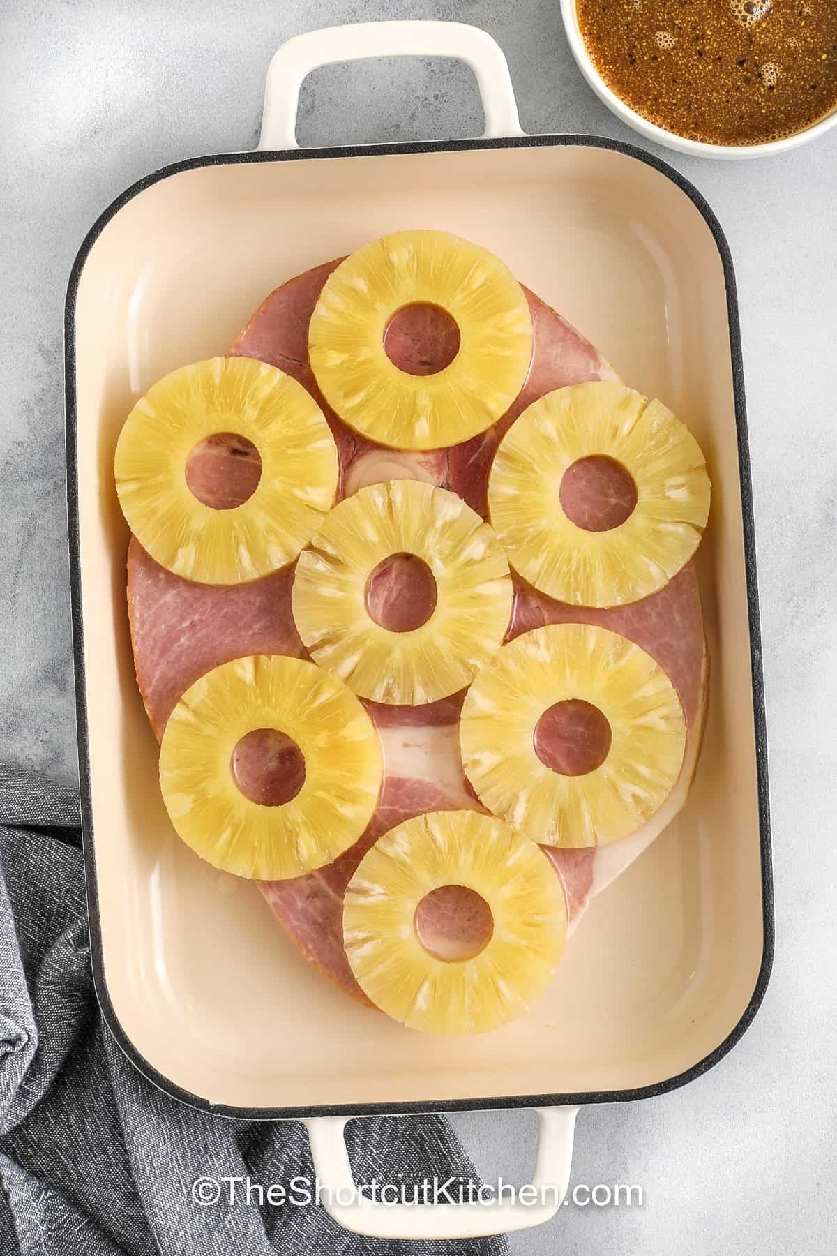 A sweet pineapple glaze turns this ham steak recipe into something extra special. The ham steak is baked in a savory sauce made with brown sugar, Dijon mustard, soy sauce, garlic, and black pepper. Pineapple provides a sweet, tangy contrast that pairs well with the ham. This dish works for weeknight dinners or casual gatherings. Serve it with roasted vegetables, mashed potatoes, or rice for an easy, flavorful, and satisfying meal for the whole family. #theshortcutkitchen adding pineapple rings to ham in dish to make Ham Steak