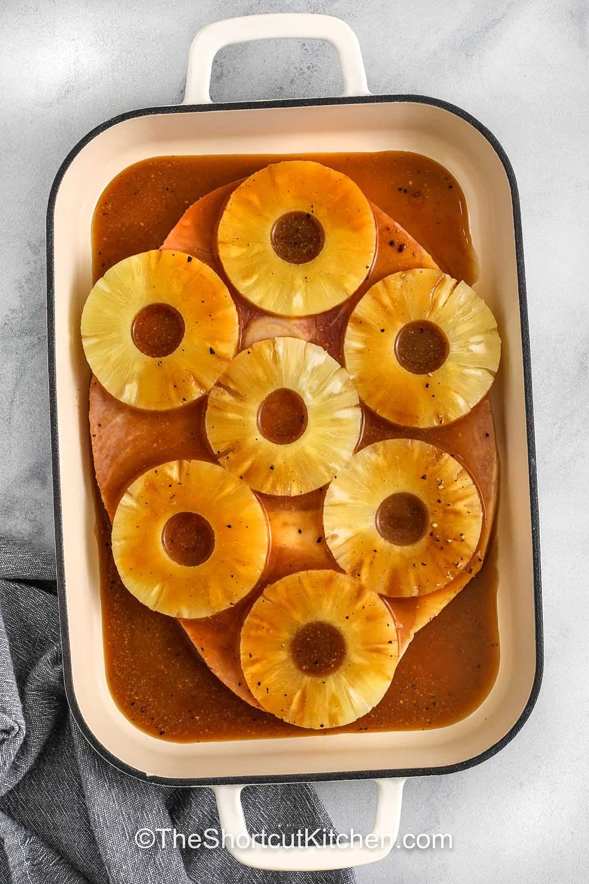 Try this ham steak recipe with pineapple and a sweet glaze for a flavorful, easy meal. The ham steak is coated in a brown sugar sauce with mustard and soy sauce, enhancing every bite. Pineapple adds brightness and helps keep the ham tender while baking. This dish is perfect for a casual family dinner or a small holiday celebration. Serve it alongside green beans, roasted potatoes, or a fresh salad to create a comforting dinner everyone will love. #theshortcutkitchen adding glaze over pineapple and ham to make a Ham Steak