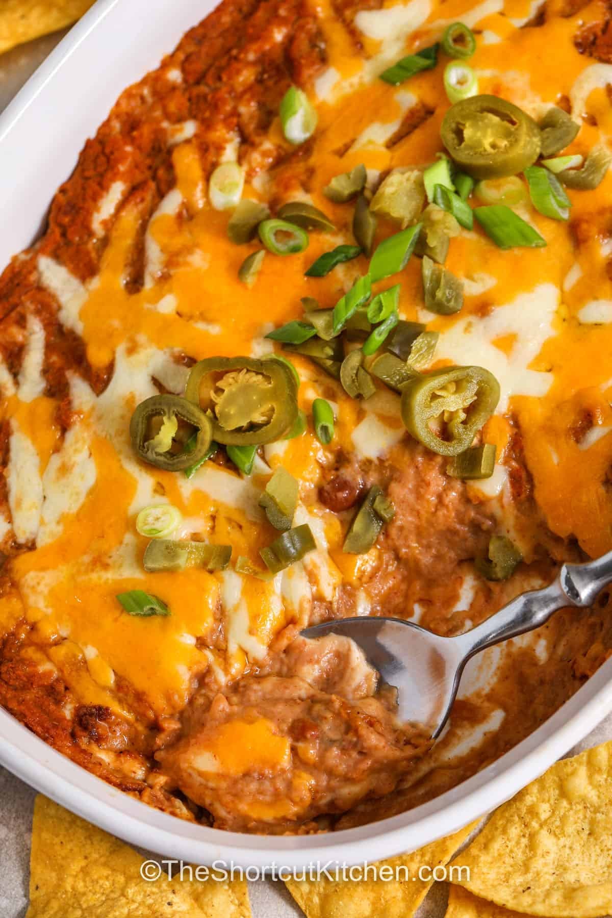 Refried bean dip is an easy hot appetizer that is always a hit at parties. This baked bean dip combines refried beans, salsa, taco seasoning, and sour cream with melted cheese for classic Tex-Mex flavor. Scoop it up with tortilla chips or crackers for a warm snack that is great for sharing. Serve this party dip at potlucks, family gatherings, or game day events when you want a simple appetizer that comes together with everyday ingredients. #theshortcutkitchen refried bean dip with a scoop taken out of it