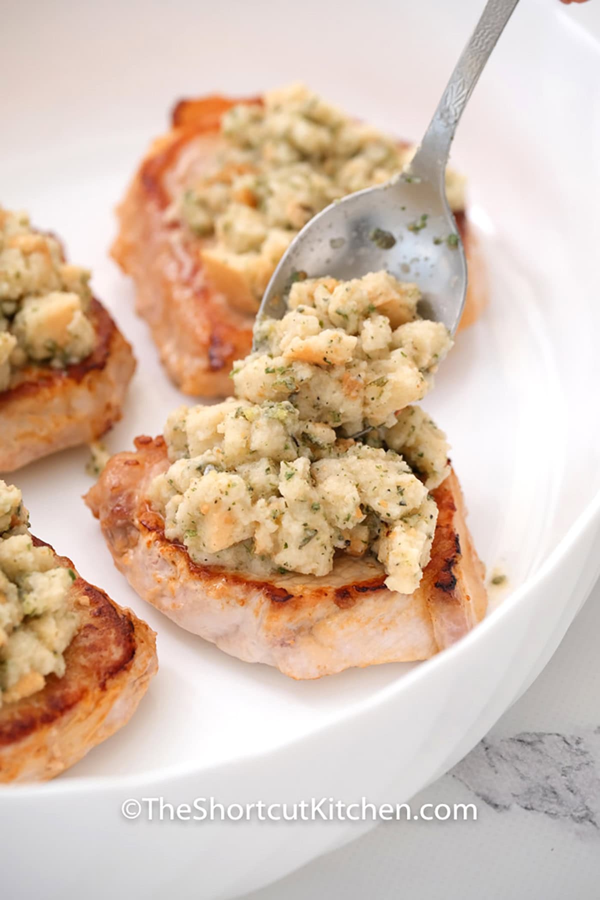 Comfort food has never been easier than this smothered pork chops recipe. By pan-searing the chops first, you lock in the flavor before smothering them in a creamy sauce and herb-seasoned stuffing. Serve with a side of green beans for a complete, hearty meal. #theshortcutkitchen adding stuffing to pork chops to make Smothered Pork Chops Recipe