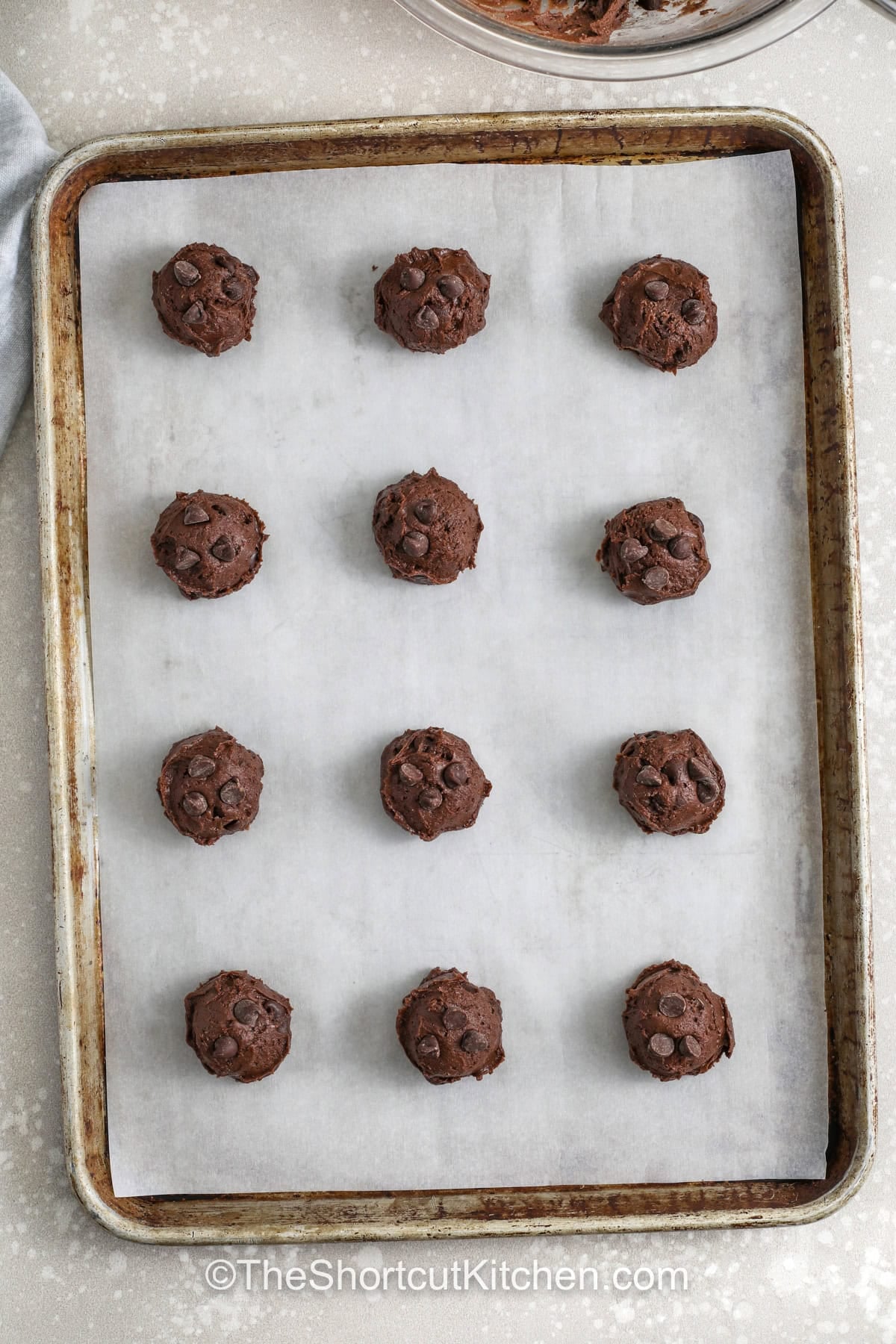 Chocolate cake cookie mix makes this recipe a foolproof way to enjoy bakery quality cookies without complicated steps or long prep time. Each cookie bakes up soft, thick, and full of chocolate flavor with just the right balance of sweetness and richness. The melted chocolate chips add extra gooey pockets that make every bite even more satisfying and indulgent. #theshortcutkitchen Chocolate Cake Mix Cookies dough rolled into a ball on a sheet pan before cooking