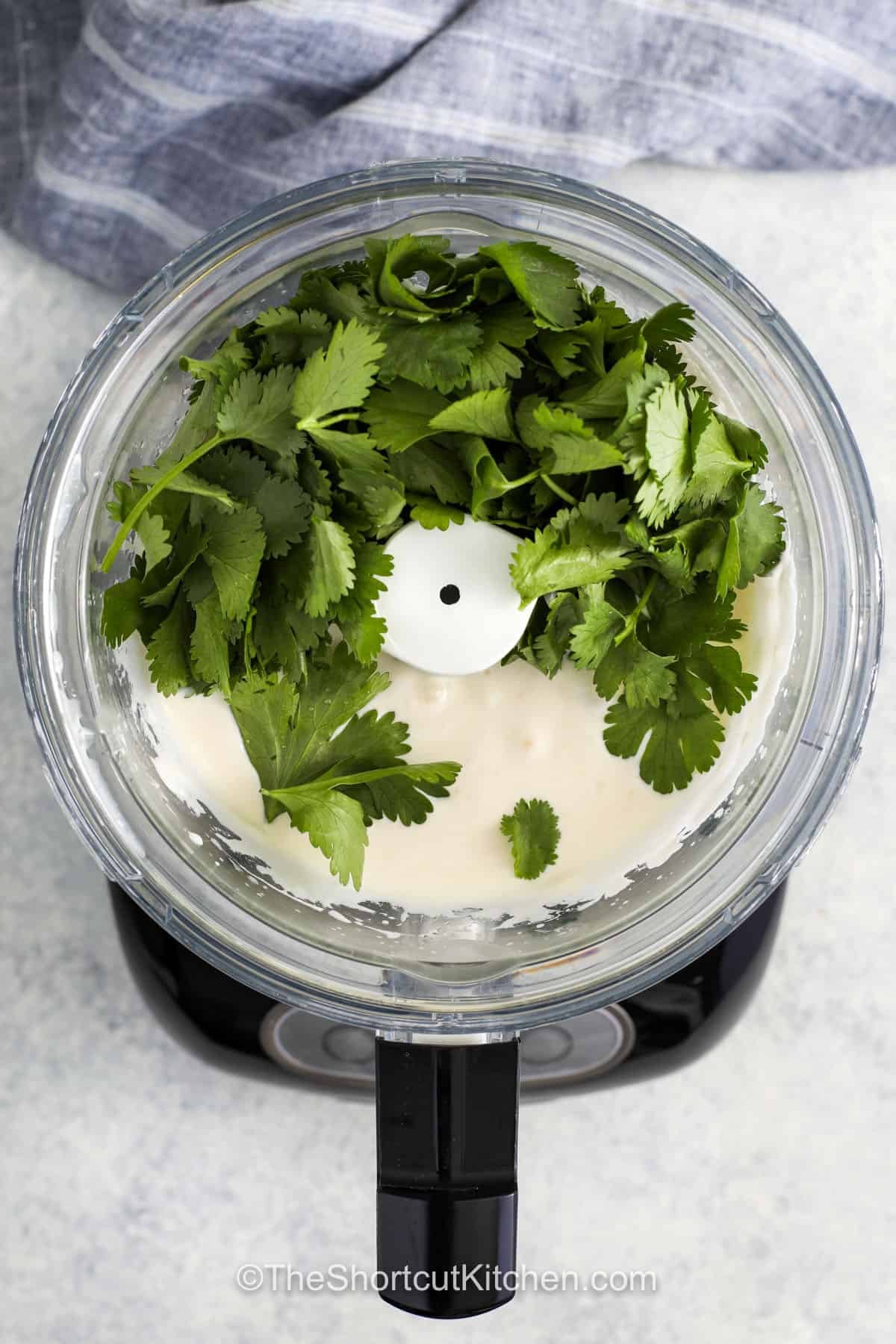 Cilantro garlic sauce brings a fresh, creamy boost to everyday meals. This easy homemade sauce blends sour cream, mayo, lime, and garlic with bright cilantro for the best flavor in every bite. It works as a dip, dressing, or drizzle for tacos, grilled chicken, roasted vegetables, and rice bowls. Keep a batch in the fridge and use it all week to add extra flavor without any extra effort. It is a simple way to prep make-ahead meals that taste super special. #theshortcutkitchen adding cilantro to ingredients in blender to make Cilantro Garlic Sauce