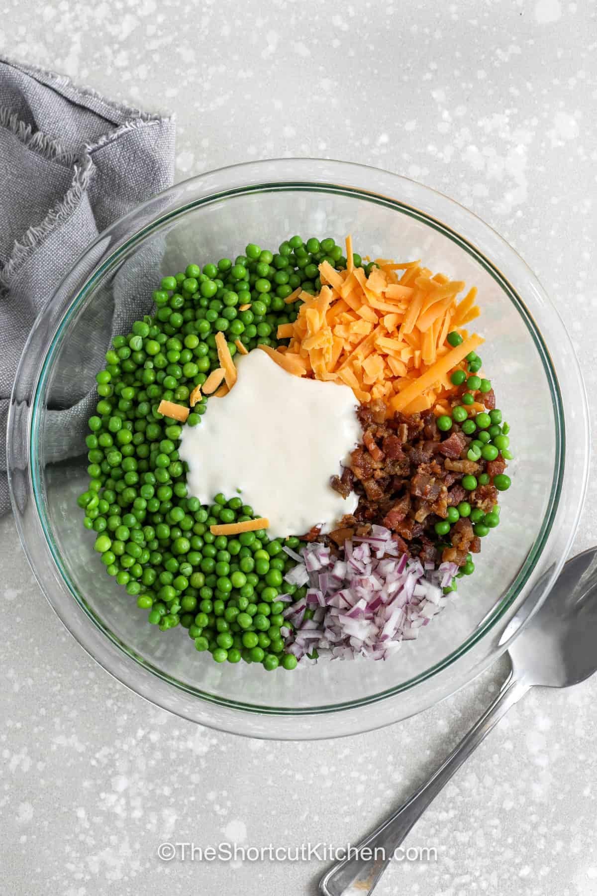 This pea salad with bacon is an easy pea salad recipe made with thawed frozen peas, sharp cheddar cheese, crispy bacon bits, and finely diced red onion in a creamy, tangy dressing. It is the kind of cold side dish that works for spring lunches, holiday dinners, and warm-weather gatherings. If you are looking for a creamy pea salad that is quick, budget-friendly, and full of texture, this one checks every box. #theshortcutkitchen adding ingredients together to make Pea Salad with Bacon