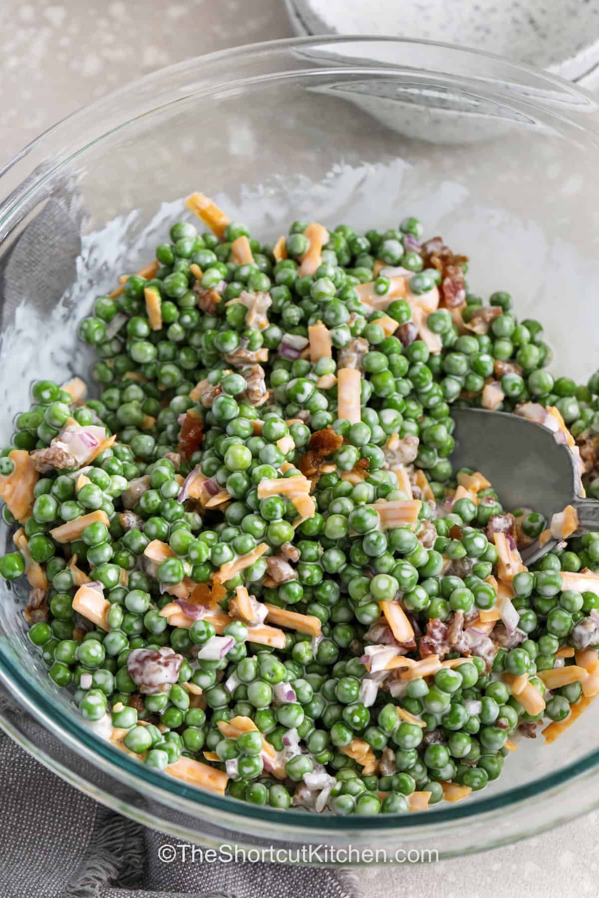 Pea salad with bacon is a creamy, classic cold side dish made with frozen peas, cheddar cheese, bacon bits, red onion, and coleslaw dressing. This easy pea salad is perfect for potlucks, BBQs, Easter dinner, and summer cookouts because it comes together fast with shortcut ingredients and can be made ahead. It is a great old-fashioned pea salad recipe when you need something simple, cool, and crowd-friendly. #theshortcutkitchen glass bowl with Pea Salad with Bacon