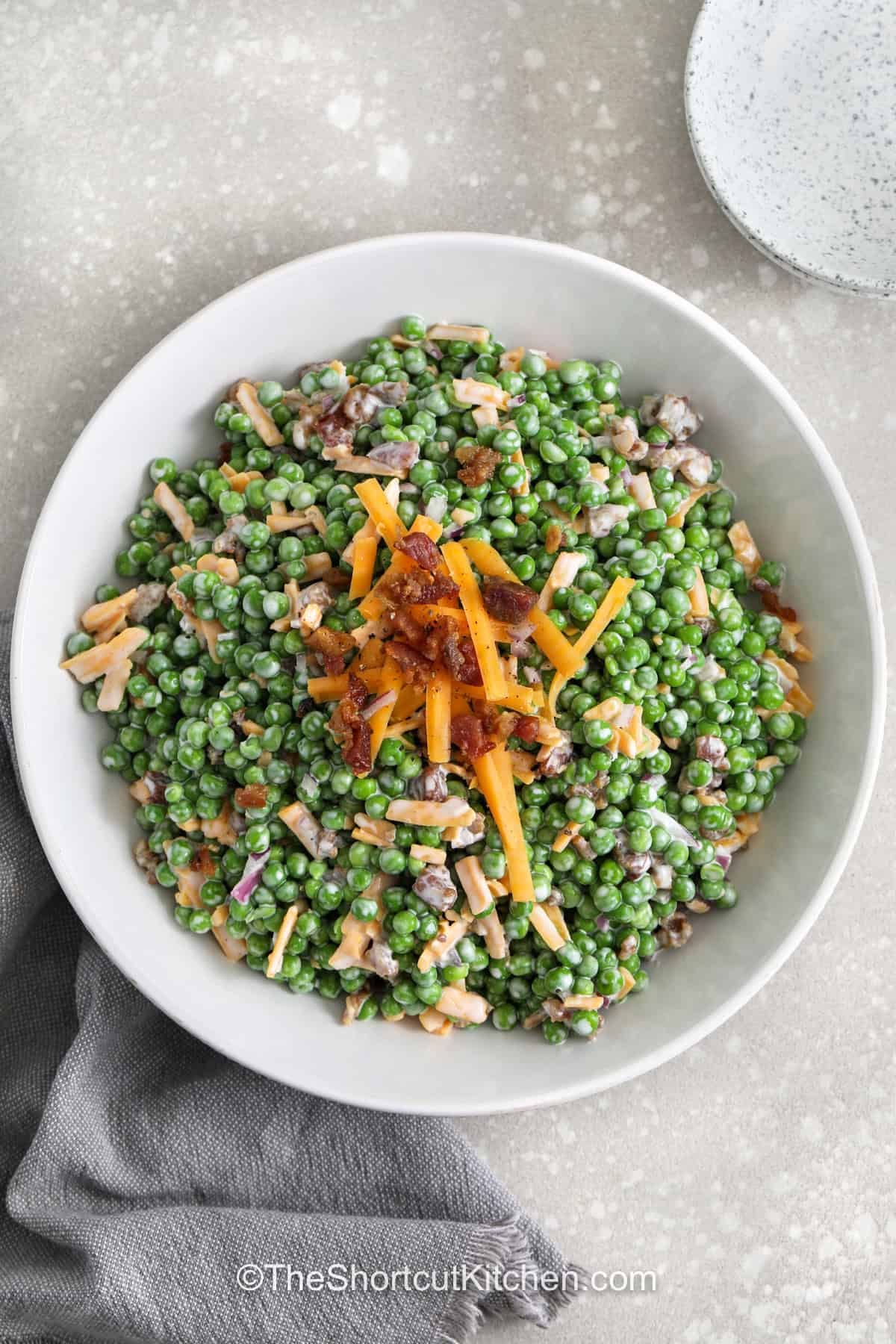 Pea salad with bacon is a creamy pea salad recipe that keeps things simple with frozen peas, prepared dressing, cheddar cheese, and bacon bits. It is fast enough for a weeknight dinner side but special enough for potlucks and holiday meals. This cold pea salad has the perfect balance of sweet peas, savory bacon, and tangy dressing, with a little crunch from fresh red onion. #theshortcutkitchen plated dish of creamy Pea Salad with Bacon