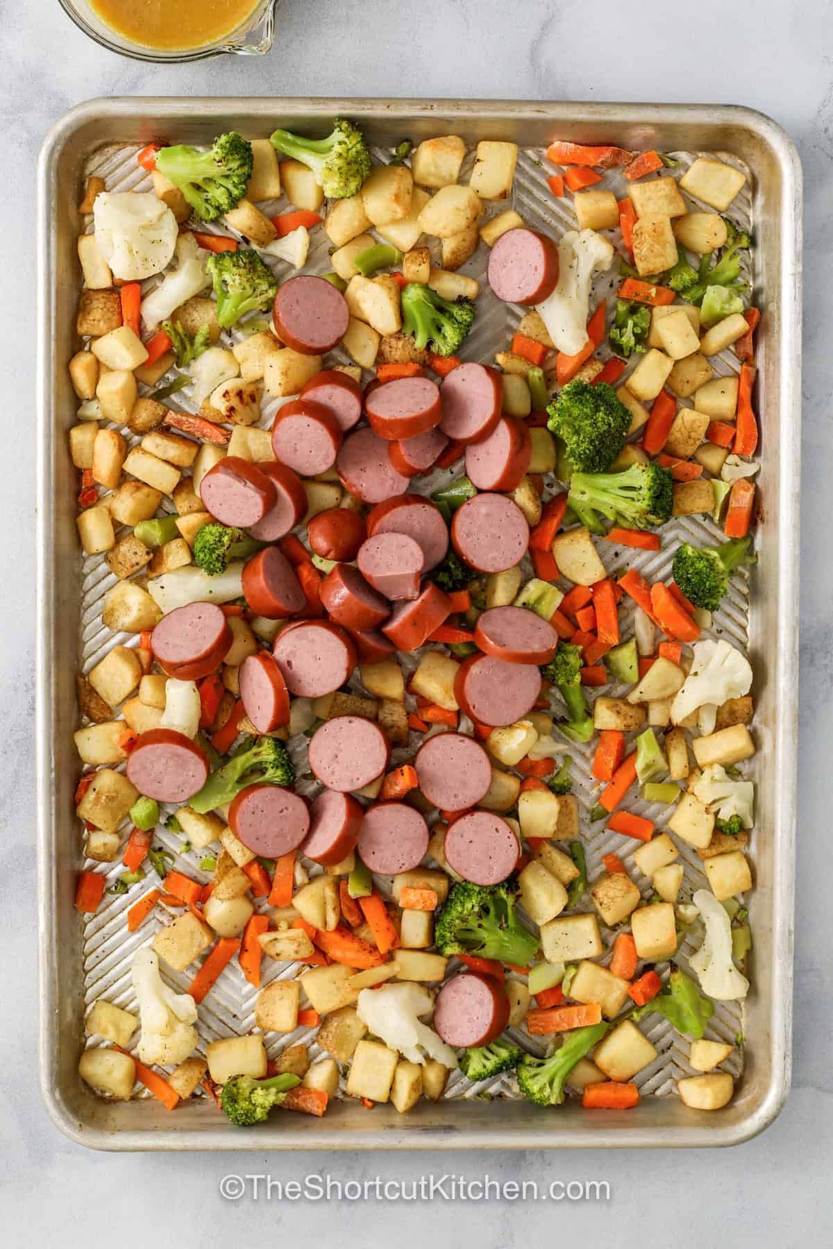 Sheet pan sausage and veggies is the kind of easy dinner everyone looks forward to. Smoked sausage, frozen vegetables, and crispy potatoes roast together in one pan for big flavor with very little cleanup. The honey Dijon glaze adds a sweet and savory finish that makes every bite extra tasty. This simple weeknight dinner is hearty, colorful, and perfect for busy evenings. Save this easy sheet pan recipe for your next family meal. #theshortcutkitchen adding sausages to hash browns and veggies to make Sheet Pan Sausage and Veggies