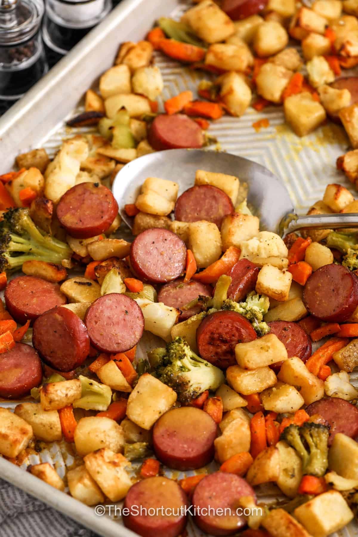 Sheet pan sausage and veggies turn simple freezer staples into a dinner everyone will love. Smoked sausage, roasted potatoes, and colorful vegetables cook together in one pan for an easy family meal. The honey Dijon glaze adds just the right amount of sweet and savory flavor. This hearty weeknight recipe is filling, comforting, and easy to prep with minimal dishes. Save this easy dinner recipe for your next busy night. #theshortcutkitchen delicious Sheet Pan Sausage and Veggies on a sheet pan with a spoon taking some off