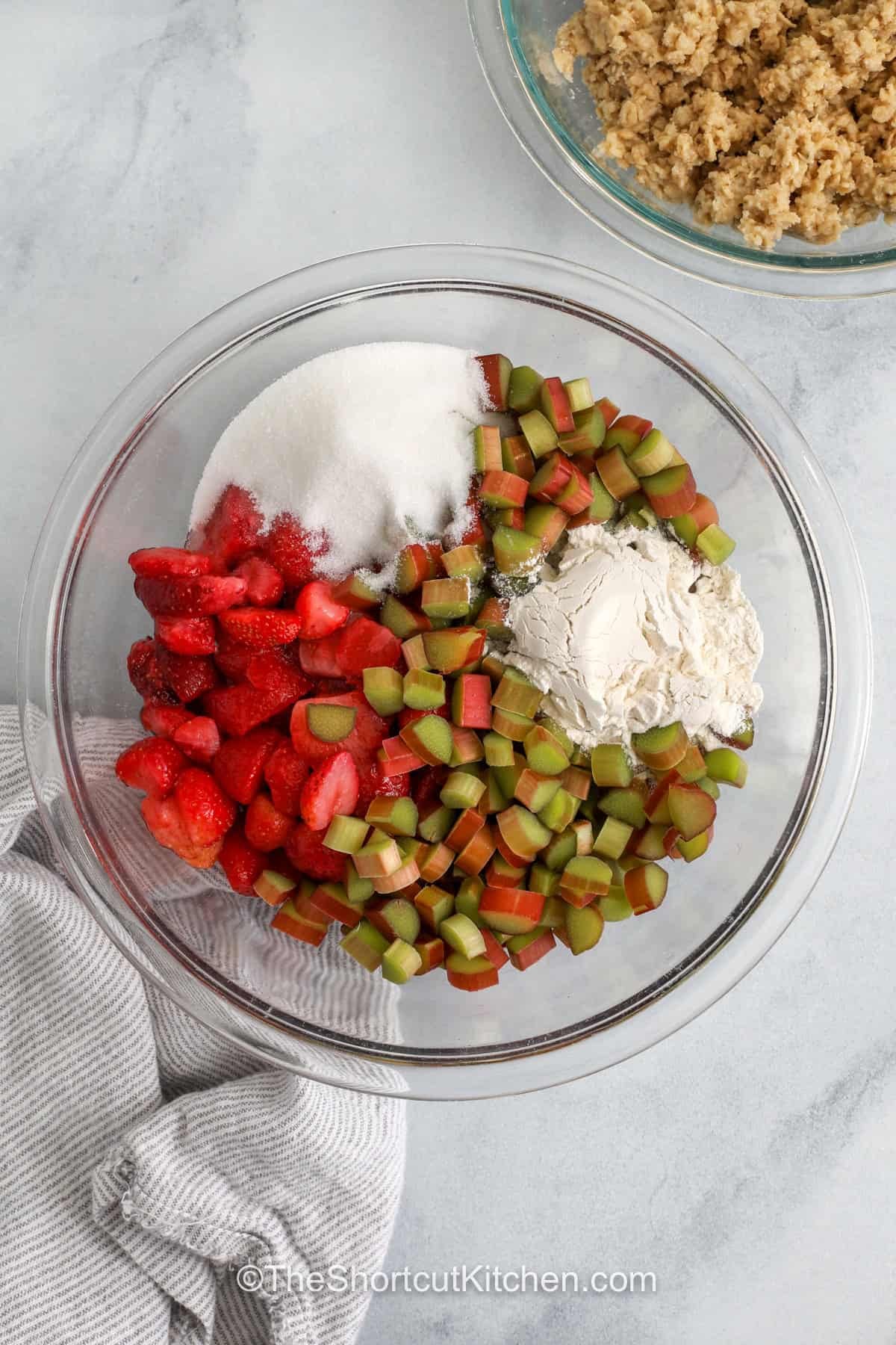 Strawberry rhubarb crisp is a classic fruit dessert that is easy to make and so tasty. Sweet strawberries mix with tart rhubarb while a buttery topping adds a nice crunch. A cookie mix keeps this recipe simple but still gives it a homemade taste. It is a great way to use fresh fruit in season or fruit from the freezer. Serve it warm for a cozy treat that everyone will want more of. #theshortcutkitchen adding flour and sugar to strawberries and rhubarb to make Strawberry Rhubarb Crisp