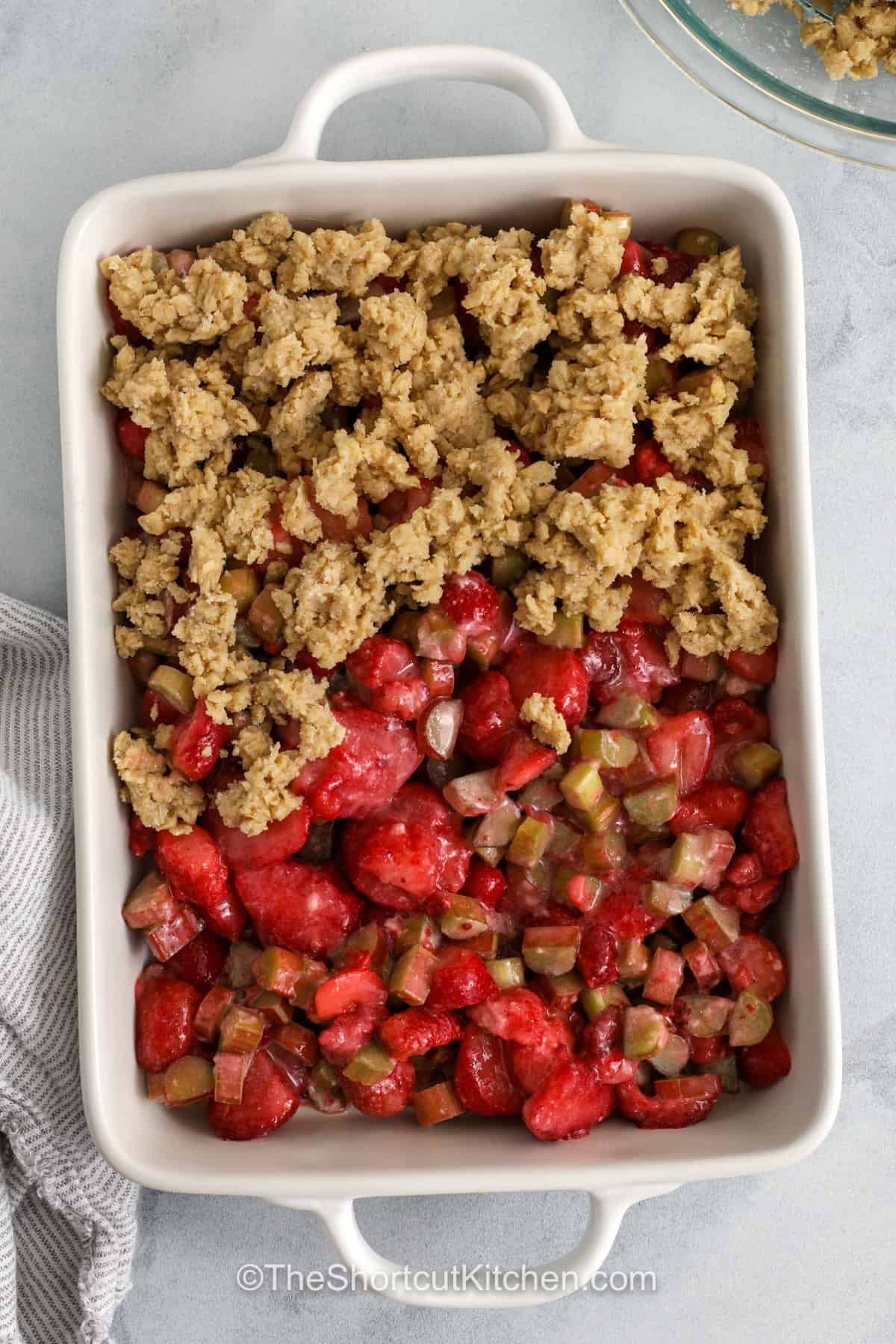 Strawberry rhubarb crisp is a classic fruit dessert that is both simple and satisfying. Sweet strawberries balance the tart rhubarb while a buttery topping adds just the right crunch. Using a cookie mix makes this recipe quick to assemble without losing that homemade taste. It is a great way to use seasonal produce or frozen fruit from your freezer. Serve it warm for a cozy treat that everyone will come back for again and again. #theshortcutkitchen adding crisp topping on top of strawberries and rhubarb to make Strawberry Rhubarb Crisp