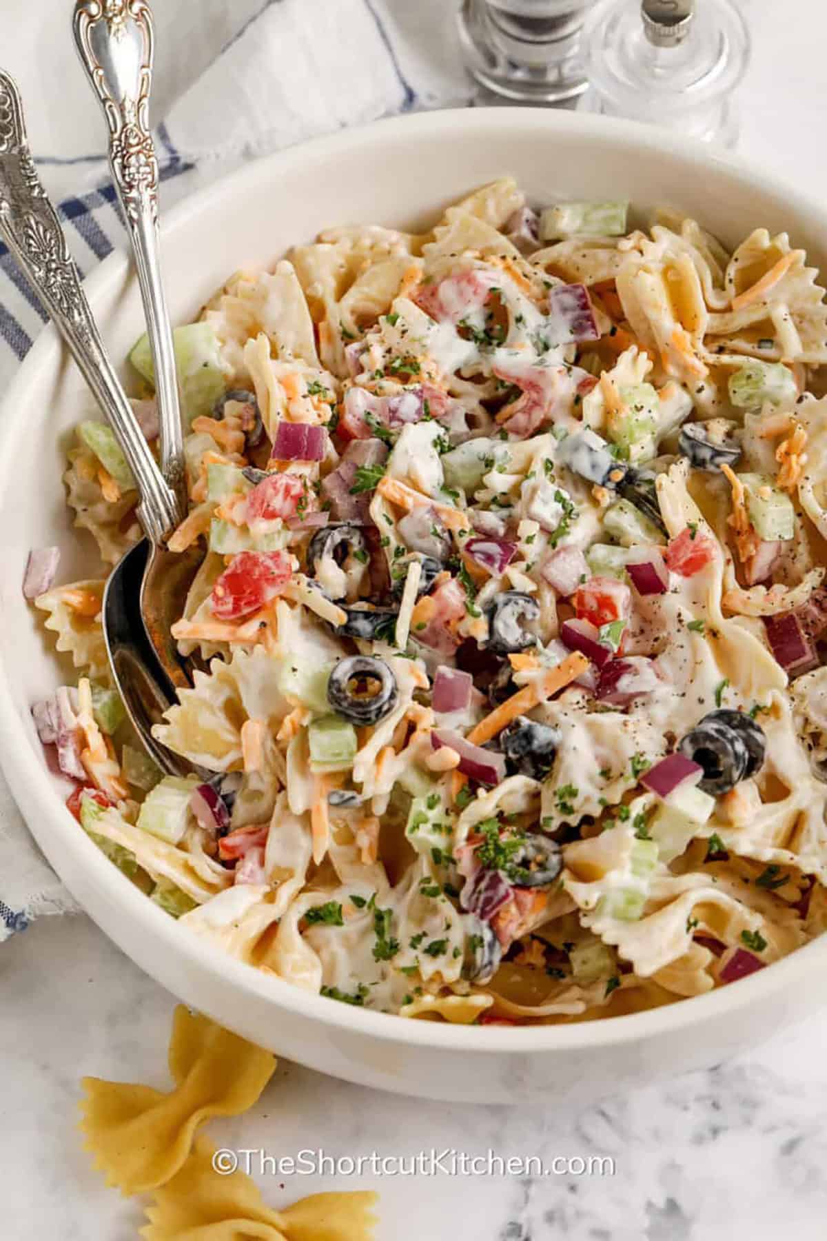 This easy bow tie pasta salad recipe is made with ranch dressing, tomatoes, celery, red onion, olives, and shredded cheddar cheese. It is a creamy pasta salad that is budget-friendly, simple to prepare, and easy to customize with bacon, chicken, peas, cucumber, or bell peppers for extra flavor. Bring this to the next potluck, BBQ, or summer picnic. #theshortcutkitchen bowl of Bow Tie Pasta Salad with a fork and spoon