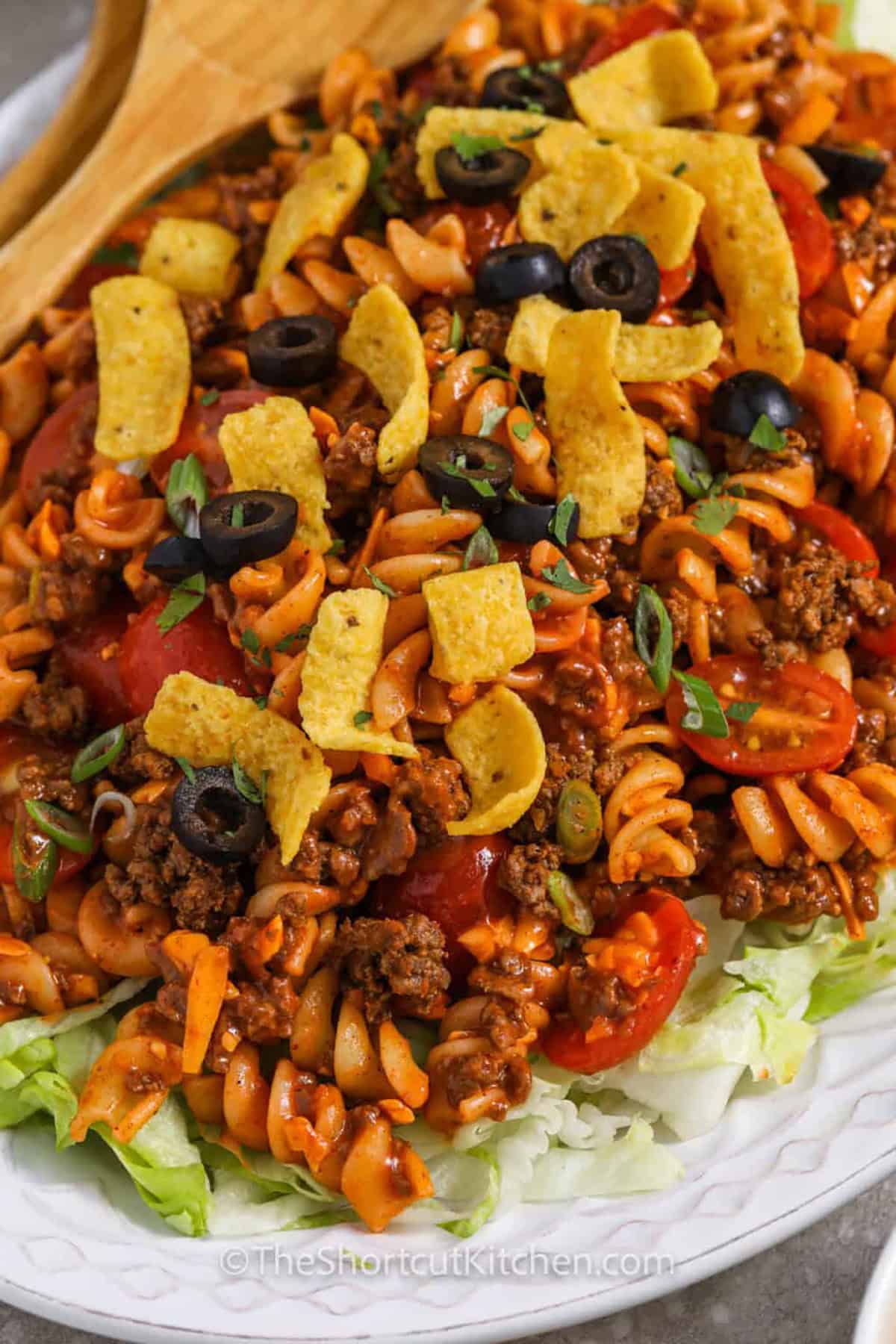 This taco pasta salad is an easy 30-minute recipe made with rotini pasta, seasoned ground beef, cherry tomatoes, cheddar cheese, shredded iceberg lettuce, Catalina dressing, and crunchy corn chips. It is a cold pasta salad with taco flavor that works as a hearty side dish or an easy dinner. #theshortcutkitchen Taco Pasta Salad on lettuce