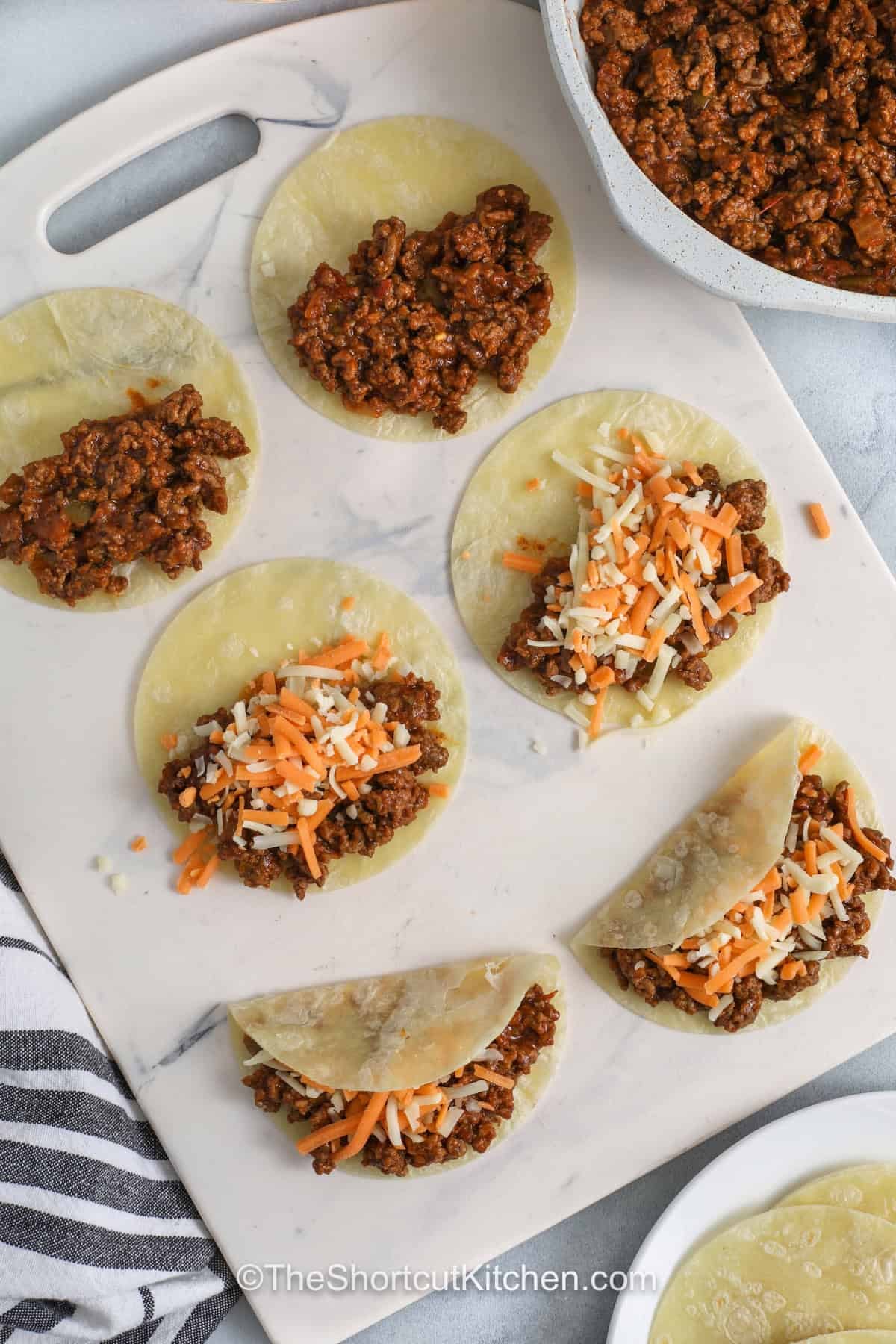 Easy baked tacos are stuffed with seasoned ground beef and shredded Mexican cheese, then pressed and baked between hot sheet pans until crisp. This recipe is simple, budget friendly, and perfect for serving with taco toppings like lettuce, tomatoes, sour cream, guacamole, pico de gallo, and jalapenos. They make a fun dinner perfect for any night. Save this easy taco night recipe. #theshortcutkitchen adding cheese and beef to tortillas to make Baked Tacos