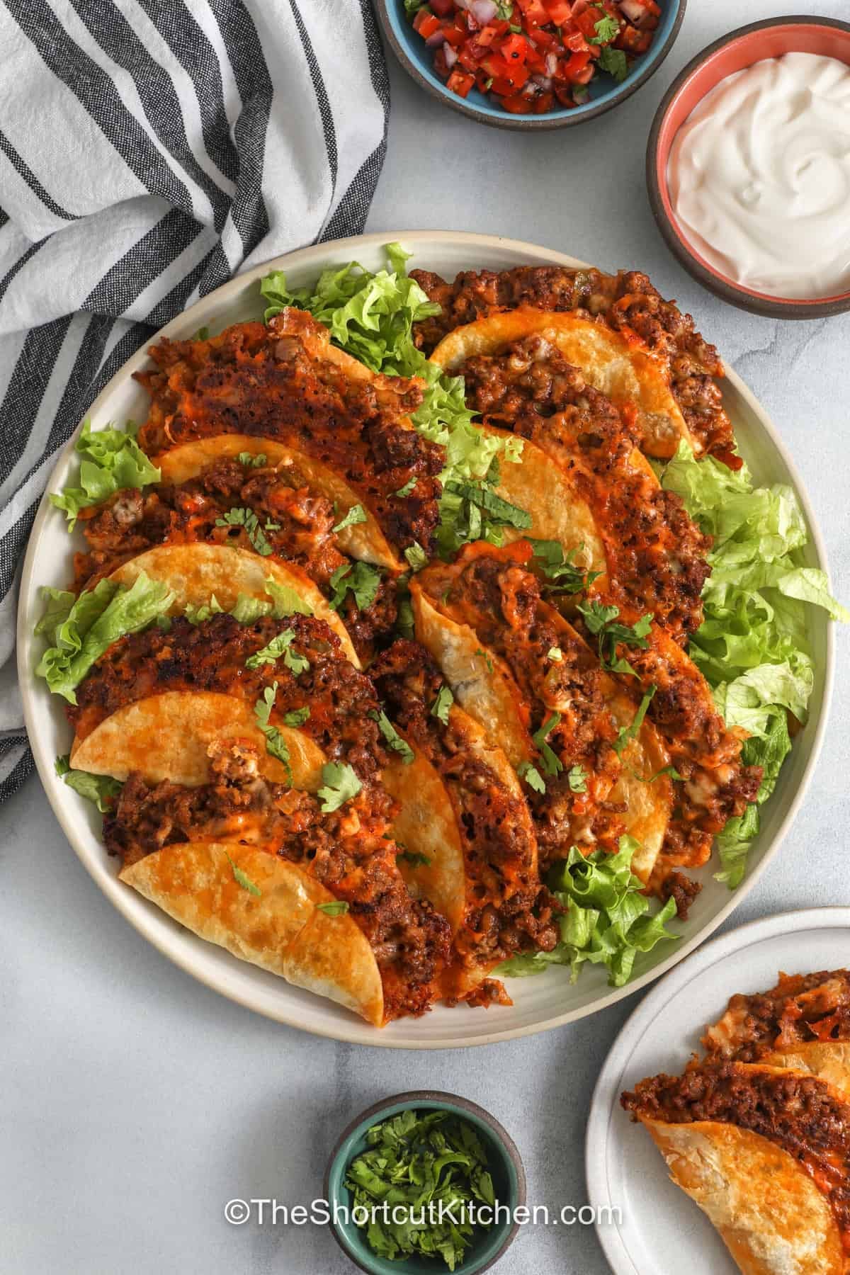 Crispy baked tacos are the perfect taco night shortcut when you want something easy, cheesy, and family friendly. Ground beef is cooked with salsa and taco seasoning, tucked into flour tortillas with cheese, and baked until golden. Serve these Baked Tacos with shredded lettuce, tomatoes, sour cream, guacamole, pico de gallo, or jalapenos. Save this recipe for a quick weeknight meal. #theshortcutkitchen crisp Baked Tacos on a plate with bowls of sour cream and pico de Gallo