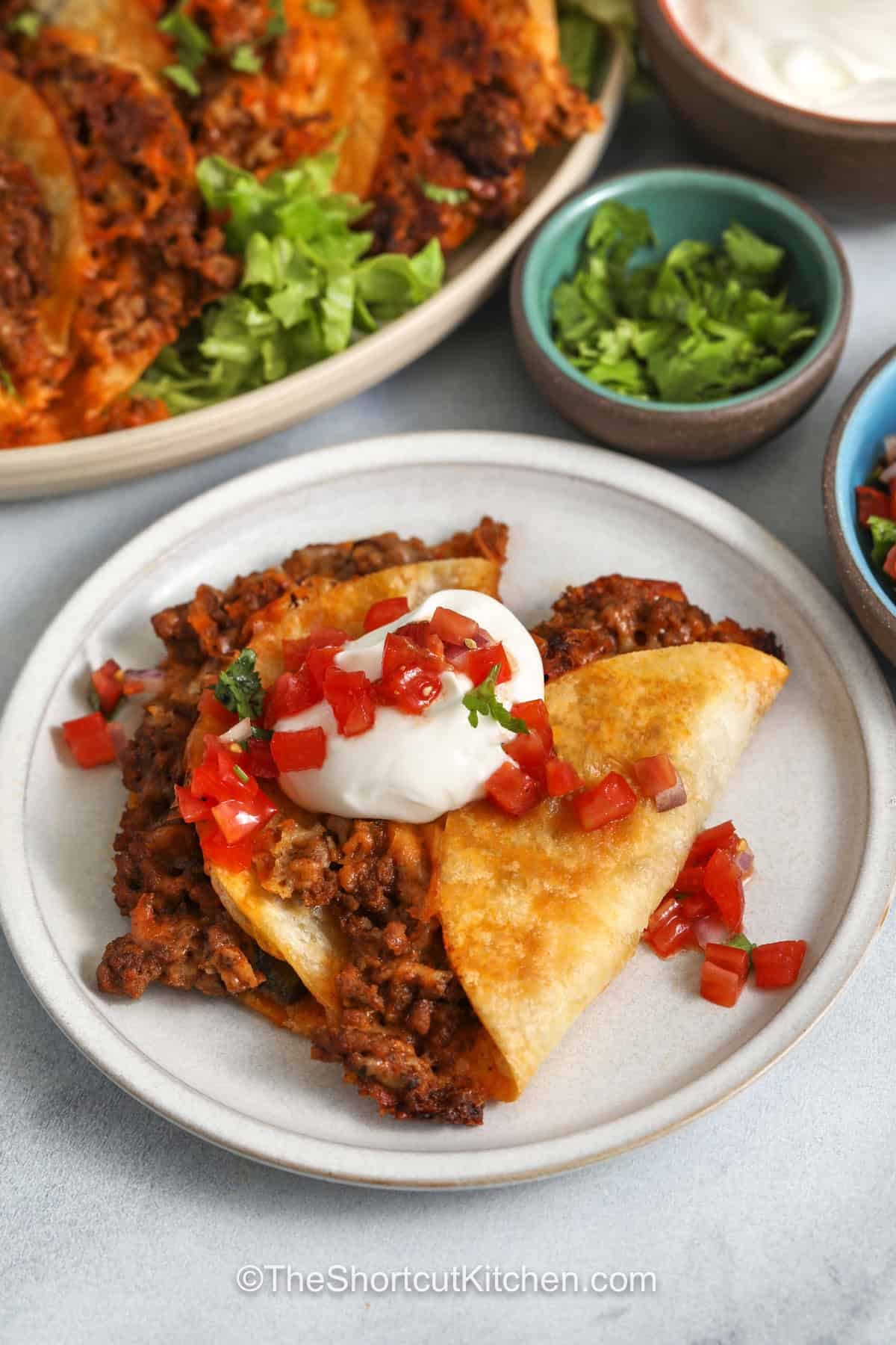 Oven baked tacos are made with flour tortillas, seasoned ground beef, salsa, and shredded Mexican cheese, then baked until crispy and golden. This easy recipe uses a hot sheet pan method for crunchy edges without frying. Add lettuce, tomatoes, sour cream, guacamole, pico de gallo, or jalapenos for a fun taco night dinner everyone can customize. Save this recipe for your next taco night. #theshortcutkitchen plated crispy Baked Tacos with sour cream and pico de Gallo