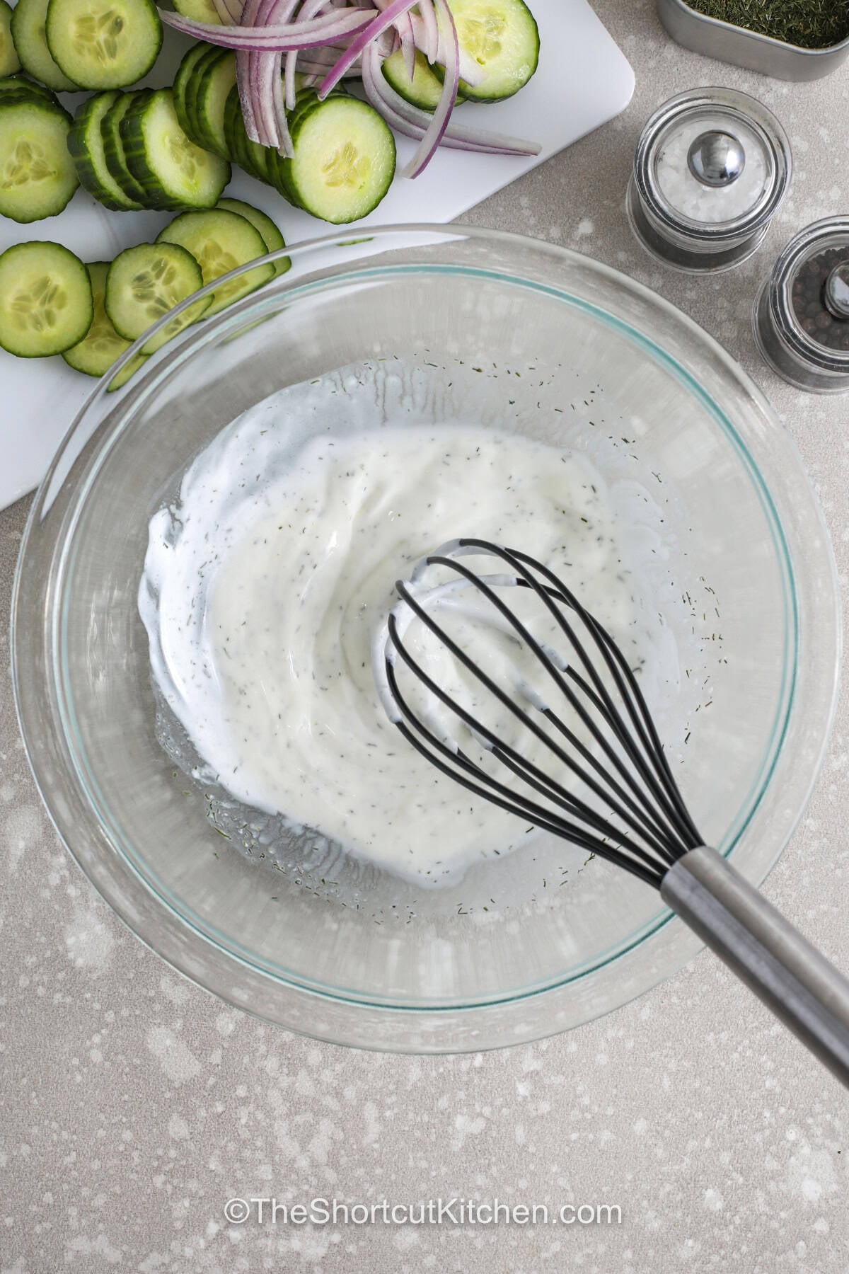 This creamy cucumber salad recipe is a chilled summer favorite made with sliced cucumbers, red onion, and tangy sour cream dressing. Every spoonful is packed with fresh dill flavor and crisp texture that tastes great beside burgers, hot dogs, or barbecue chicken. It is an easy make-ahead salad for picnics, potlucks, and family dinners during warm weather months. Make this easy cucumber salad tonight. #theshortcutkitchen mixing Creamy Cucumber Salad Recipe sauce in a bowl