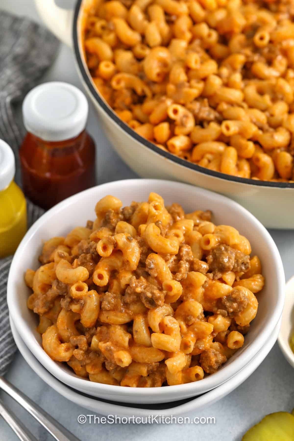 This Cheeseburger Macaroni recipe is a creamy one-pot pasta dinner made with lean ground beef, cheddar cheese, tomato sauce, ketchup, and mustard. The pasta cooks right in the sauce for extra flavor, then everything gets stirred with cheese for a rich, cheesy dinner. Add chopped pickles, diced tomatoes, or shredded lettuce for a burger-style finish. Pin it now for your weeknight dinners! #theshortcutkitchen bowl of One Pot CheeseBurger Pasta with pot full in the back