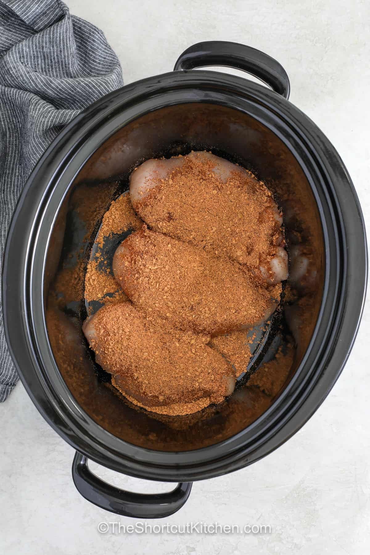 This fiesta chicken recipe transforms simple pantry staples into a creamy Tex Mex meal the whole family will love. Tender chicken cooks with salsa, taco seasoning, corn, and black beans before cream cheese is stirred in for a rich, creamy finish. Serve this Slow Cooker Fiesta Chicken in tacos, burrito bowls, wraps, or with tortilla chips for an easy dinner everyone will enjoy. Save this family favorite for your next meal. #theshortcutkitchen adding seasoning to chicken to make Fiesta Chicken Recipe