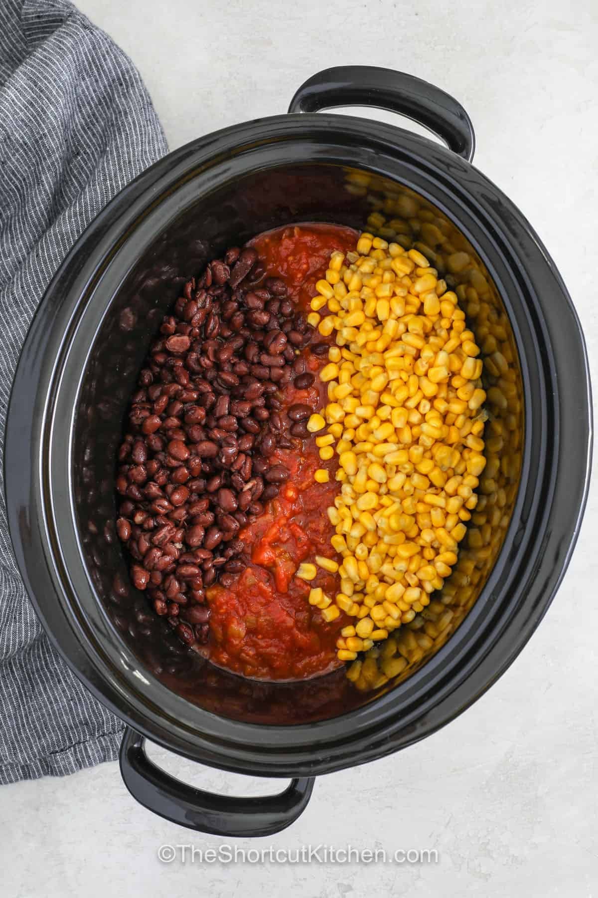Our fiesta chicken recipe turns everyday ingredients into a creamy Tex Mex dinner everyone loves. Chicken breasts cook with salsa, corn, black beans, and taco seasoning until they are tender enough to shred. Cream cheese blends into the sauce for a rich finish that is perfect for tacos, wraps, burrito bowls, or chips. This crockpot fiesta chicken is easy to serve any night of the week. Give this family favorite a place on your table. #theshortcutkitchen adding salsa , corn , beans to slow cooker to make Fiesta Chicken Recipe