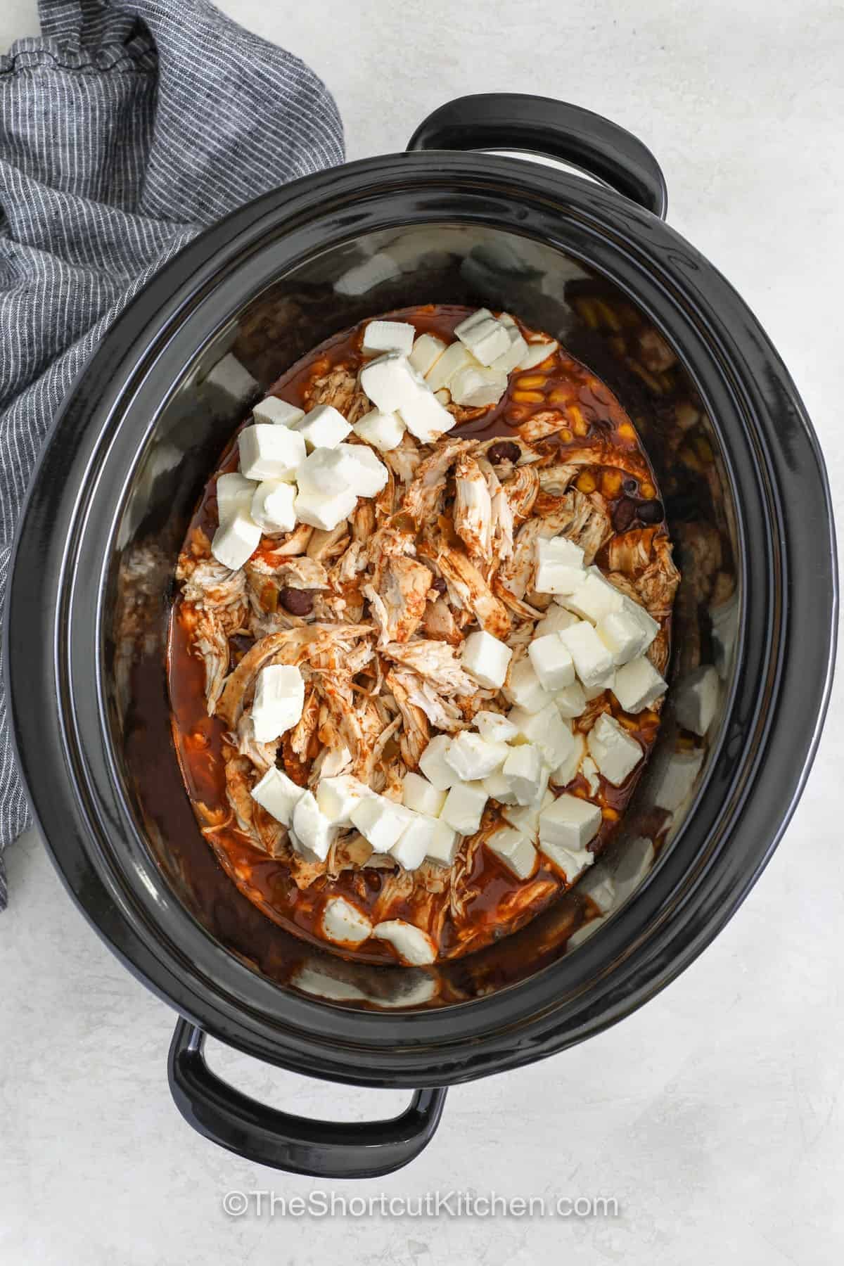 Make this fiesta chicken recipe when you need a comforting meal with very little effort. Slow Cooker Fiesta Chicken combines juicy chicken, salsa, black beans, corn, and taco seasoning into a hearty favorite. Cream cheese creates a creamy fiesta chicken that is perfect for tortillas, nachos, or rice bowls. It is a great way to feed the whole family with simple pantry ingredients. Try this recipe and make it part of your weekly menu. #theshortcutkitchen adding cream cheese and shredding chicken in slow cooker to make Fiesta Chicken Recipe