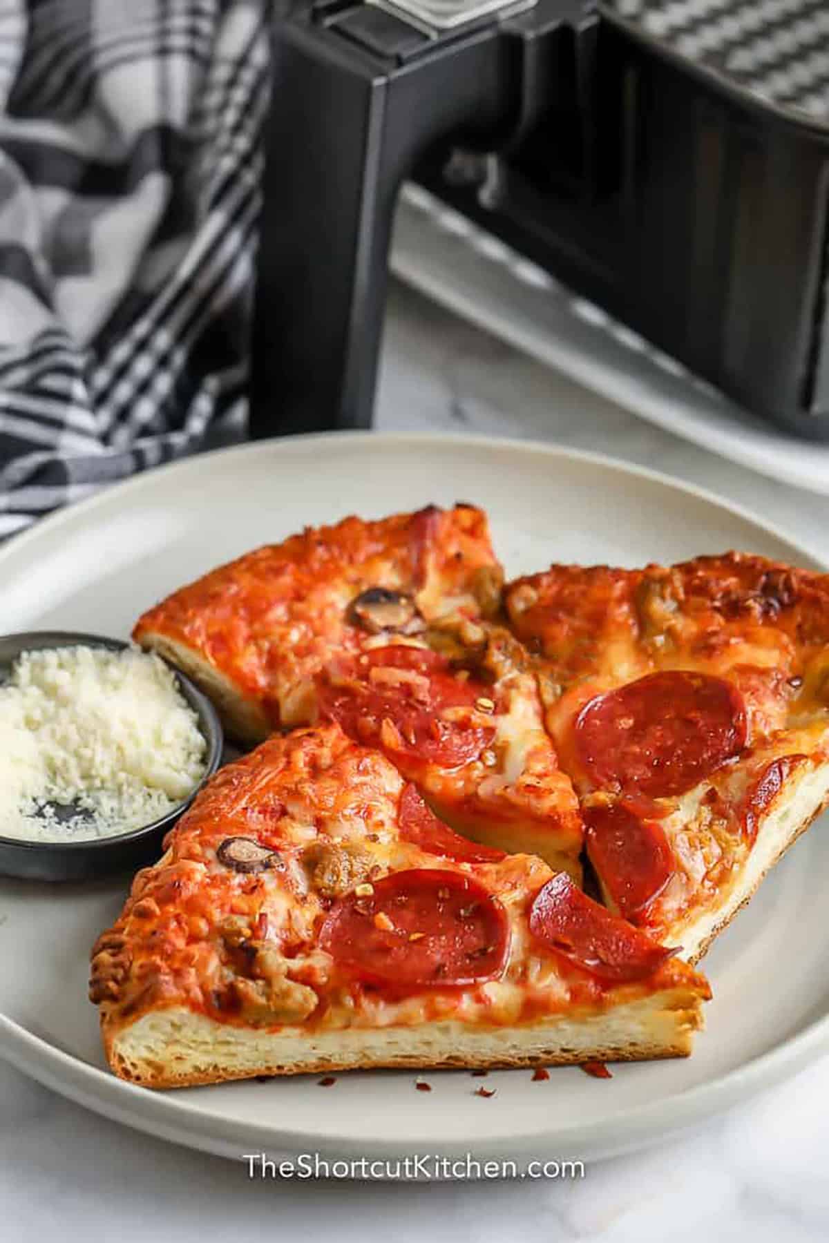 Reheat pizza in air fryer for a quick and crispy way to enjoy leftover pizza slices. The air fryer helps the crust crisp up while the cheese melts and the toppings get hot. This method works with takeout pizza, homemade pizza, thin crust pizza, or thick crust pizza. It is an easy kitchen shortcut for turning cold leftovers into a warm, cheesy snack in minutes. Pin this easy air fryer tip for your next leftover slice. #theshortcutkitchen slices of pizza on a plate with air fryer in the background to show How to Reheat Pizza in the Air Fryer