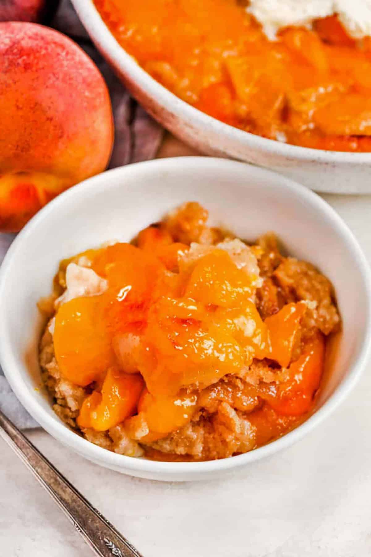 Bisquick peach cobbler is an easy baked peach dessert that uses canned peaches for quick prep and reliable flavor. The cinnamon Bisquick batter bakes around the fruit for a warm cobbler with soft texture and crisp edges. Serve this peach cobbler with canned peaches slightly warm with ice cream for the best cozy dessert. Pin this recipe so it is ready when dessert cravings hit. #theshortcutkitchen Bisquick Peach Cobbler in a bowl with dish full in the back