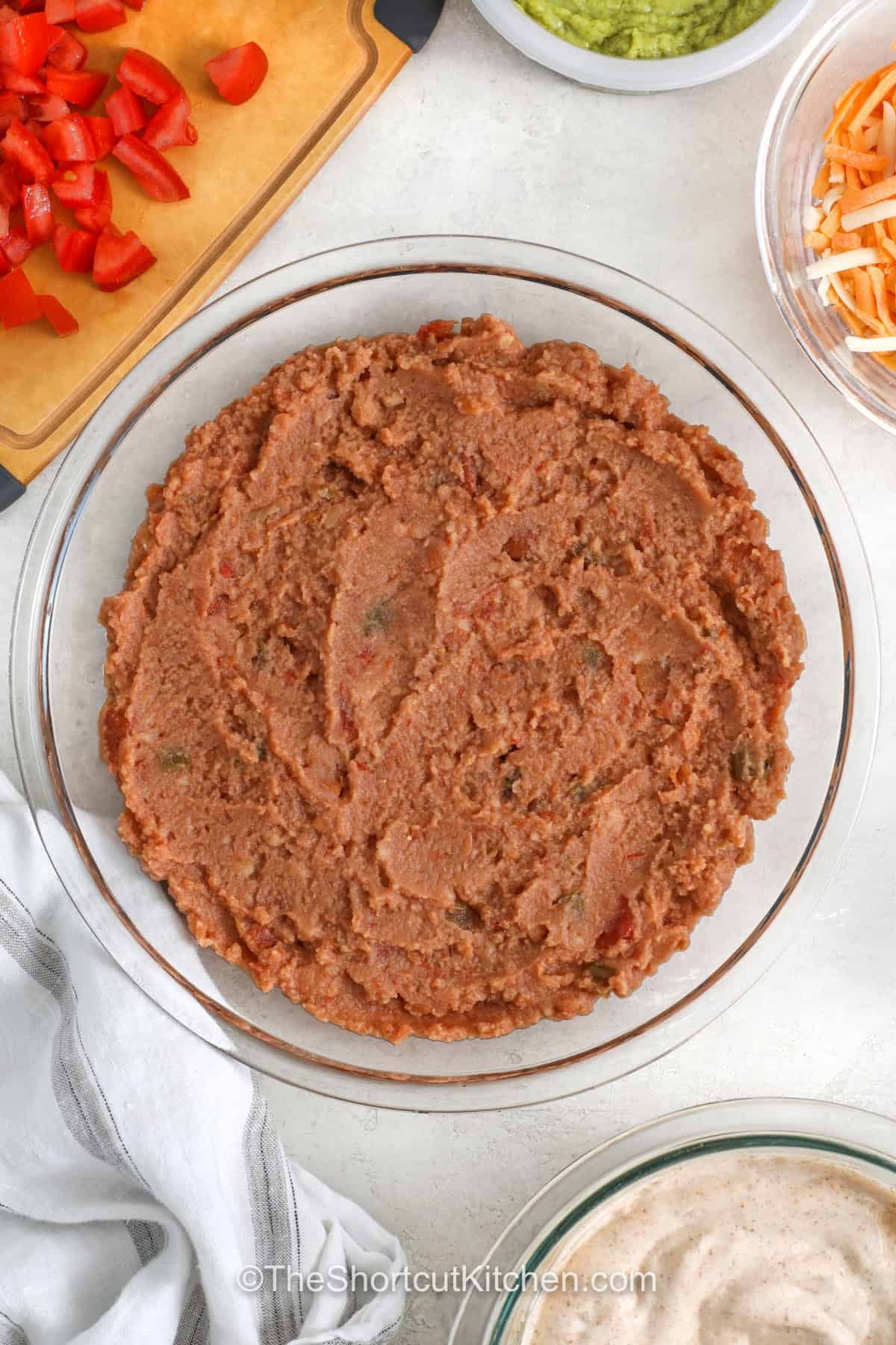5 layer dip belongs on every party menu because it is packed with colorful layers and bold flavor. Creamy beans, salsa, sour cream, cheese, and fresh toppings create an appetizer that is perfect with crunchy tortilla chips. It is easy to share and always disappears quickly at gatherings. Make this crowd favorite for your next celebration and see why everyone loves it. #theshortcutkitchen adding refried beans to dish to make 5 Layer Dip