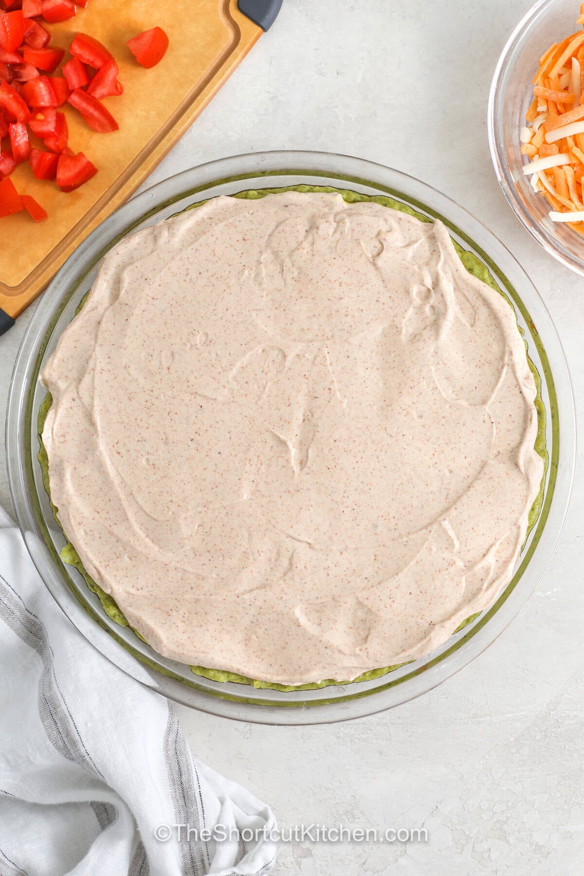 5 layer dip is the kind of appetizer everyone gathers around at parties, game days, and family get-togethers. Creamy beans, tangy sour cream, fresh salsa, shredded cheese, and crisp toppings make every bite colorful and satisfying. Serve it with tortilla chips for a crowd-pleasing snack that is always one of the first dishes to disappear. Add this easy party dip to your next menu and watch everyone come back for more. #theshortcutkitchen adding sour cream and taco seasoning layer to dish to make 5 Layer Dip