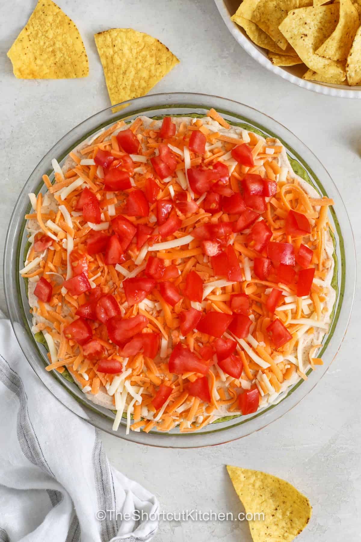 5 layer dip is always a welcome addition to any snack table or party spread. Fresh layers of beans, salsa, sour cream, cheese, and toppings create a flavorful appetizer that is easy to share. Serve it with tortilla chips for game day, potlucks, holiday parties, or family nights when everyone wants something delicious. Put this recipe on your menu and make it for your next gathering. #theshortcutkitchen adding cheese and tomatoes to dish to make 5 Layer Dip