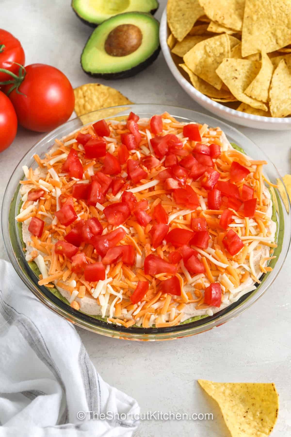 5 layer dip turns simple ingredients into a party appetizer that everyone looks forward to. The creamy layers, fresh toppings, and cheesy finish make every bite satisfying with crunchy tortilla chips. It is a great make-ahead appetizer for game day, holidays, or weekend gatherings with friends and family. Make this colorful favorite for your next event and enjoy every scoop. #theshortcutkitchen dish of 5 Layer Dip