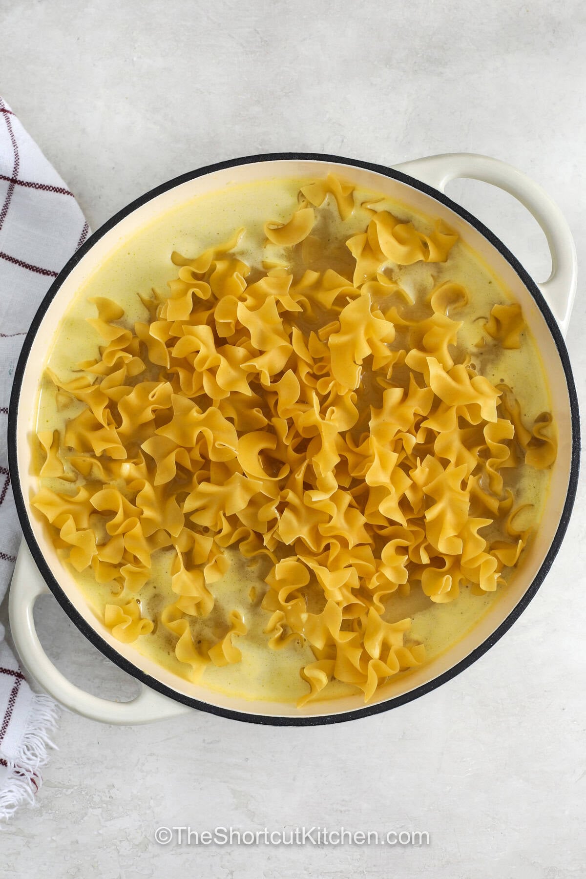 Cafeteria noodles are a creamy, buttery side dish that brings classic comfort food flavor to your table. These tender egg noodles cook in a savory chicken broth mixture for a simple recipe that pairs perfectly with chicken, turkey, pork, or meatloaf. Whether you are planning a family dinner or looking for an easy side dish, this cozy recipe is sure to please. Make these comforting cafeteria noodles today and enjoy every delicious bite. #theshortcutkitchen adding noodles to broth sauce to make Cafeteria Noodles