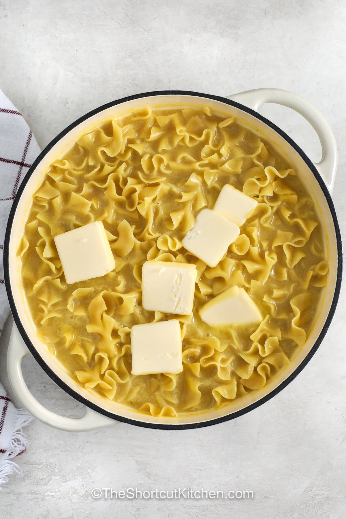 Creamy cafeteria noodles are a simple way to add something special to your next family dinner. These tender egg noodles are cooked with savory chicken broth and butter for a rich and comforting side dish. They are perfect for serving with hearty meals, holiday dinners, or whenever you want a classic recipe that feels homemade. Save this easy recipe and make these flavorful cafeteria noodles today. #theshortcutkitchen adding butter to pot to make Cafeteria Noodles