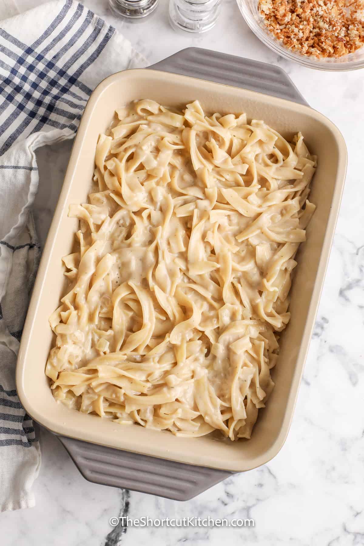 Creamy noodles and crispy onions make this French onion casserole an easy comfort food favorite. Egg noodles bake in a rich beef broth and French onion dip sauce, while a buttery Parmesan and Panko topping adds a satisfying crunch. Add chicken, ground beef, mushrooms, broccoli, or leftover vegetables to make it your own. Save this oven-baked French onion pasta recipe for busy nights. #theshortcutkitchen adding noodles with sauce to casserole dish to make French Onion Casserole