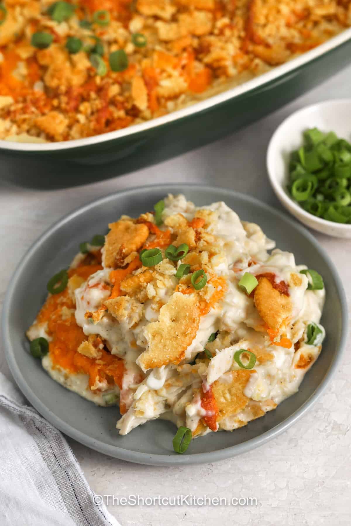 Million dollar chicken casserole is packed with the creamy, cheesy flavors that make casseroles so comforting. This delicious recipe uses simple ingredients to create a warm and satisfying meal with a golden buttery topping. Whether you use leftover chicken or rotisserie chicken, it is an easy way to make dinner feel extra special. Try this recipe tonight and share it with your family. #theshortcutkitchen Million Dollar Chicken Casserole on a plate with casserole dish full in the back
