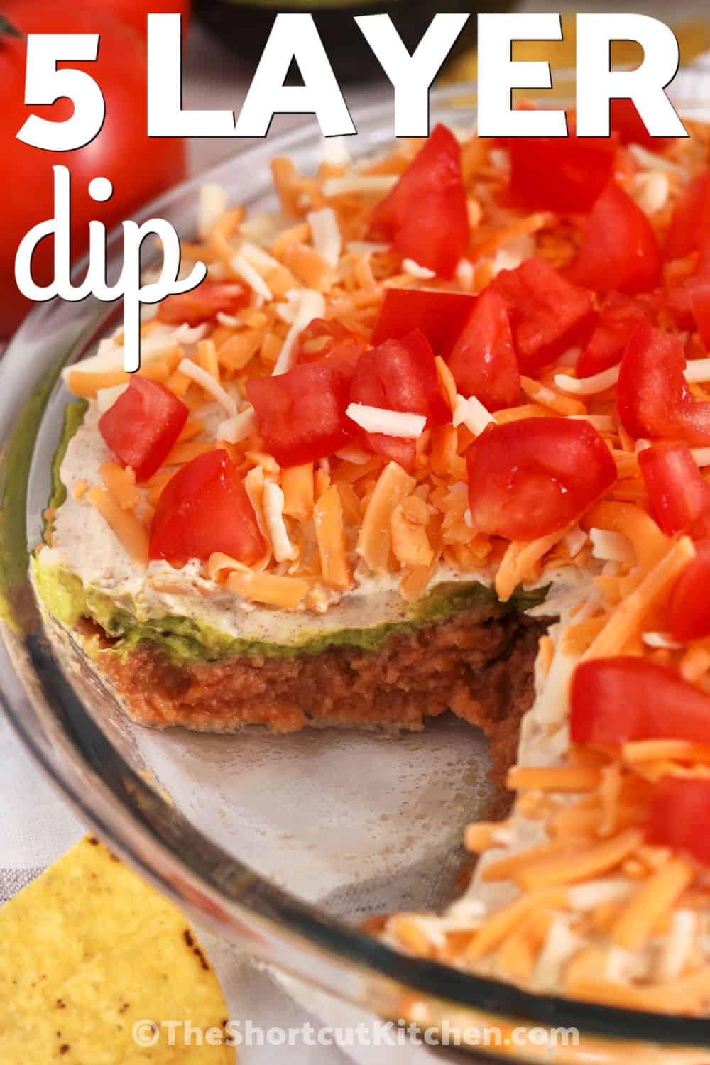 5 layer dip is the kind of appetizer everyone gathers around at parties, game days, and family get-togethers. Creamy beans, tangy sour cream, fresh salsa, shredded cheese, and crisp toppings make every bite colorful and satisfying. Serve it with tortilla chips for a crowd-pleasing snack that is always one of the first dishes to disappear. Add this easy party dip to your next menu and watch everyone come back for more. #theshortcutkitchen close up of 5 Layer Dip with a portion taken out to show the layers and a title