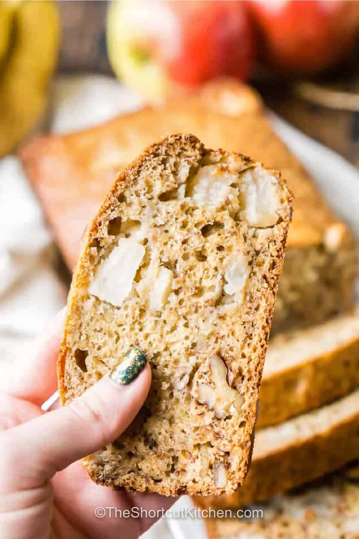 Turn ripe bananas and one fresh apple into this moist apple banana bread recipe. The easy quick bread batter is made with mashed banana, Greek yogurt, brown sugar, chopped Cortland apple, and crunchy walnuts for plenty of flavor and texture. It is a cozy homemade loaf with just 10 minutes of prep and no complicated baking techniques. Make it for breakfast, brunch, or an easy snack. Pin this banana bread with apples to bake later. #theshortcutkitchen holding a slice of Apple Banana Bread