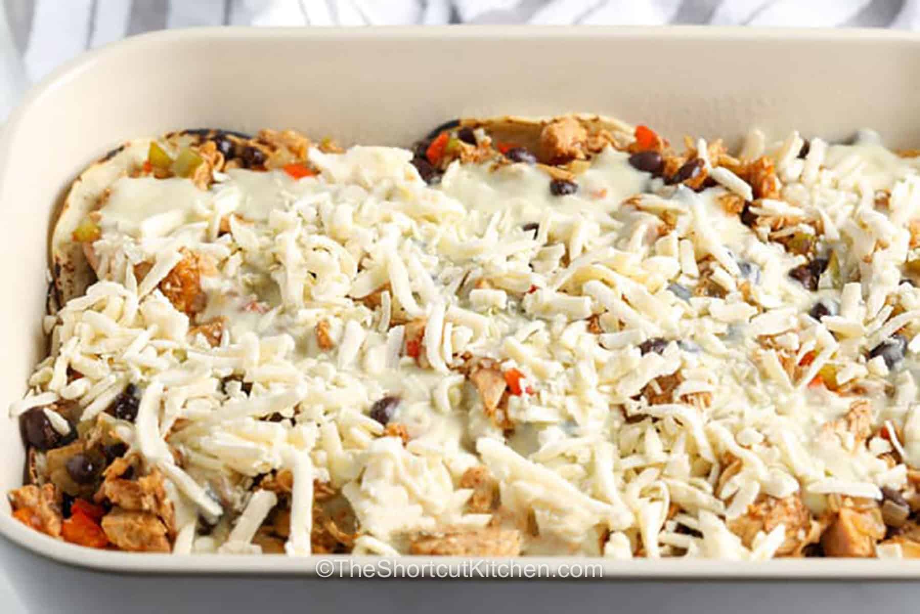 This easy chicken enchilada casserole combines shredded chicken, green enchilada sauce, corn tortillas, Monterey Jack cheese, and a creamy sauce for an easy baked dinner. Skip rolling enchiladas and enjoy all the same flavors in one comforting casserole. Perfect for busy weeknights, meal prep, and freezer meals. Save this recipe for your next family dinner. #theshortcutkitchen Chicken Enchilada Casserole in a dish before cooking.