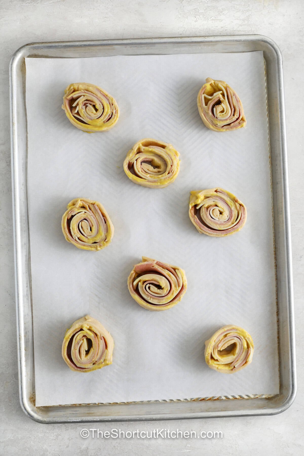 Ham and cheese pinwheels are an easy appetizer made with crescent dough, savory ham, Swiss cheese, and honey mustard. Each bite bakes up golden and melty, making these pinwheels perfect for parties, game day, lunch, or an afternoon snack. Garlic butter adds even more flavor to this simple recipe. Serve them warm and watch them disappear from the plate. Try these easy ham and cheese pinwheels today. #theshortcutkitchen Ham and Cheese Pinwheels on a sheet pan before baking