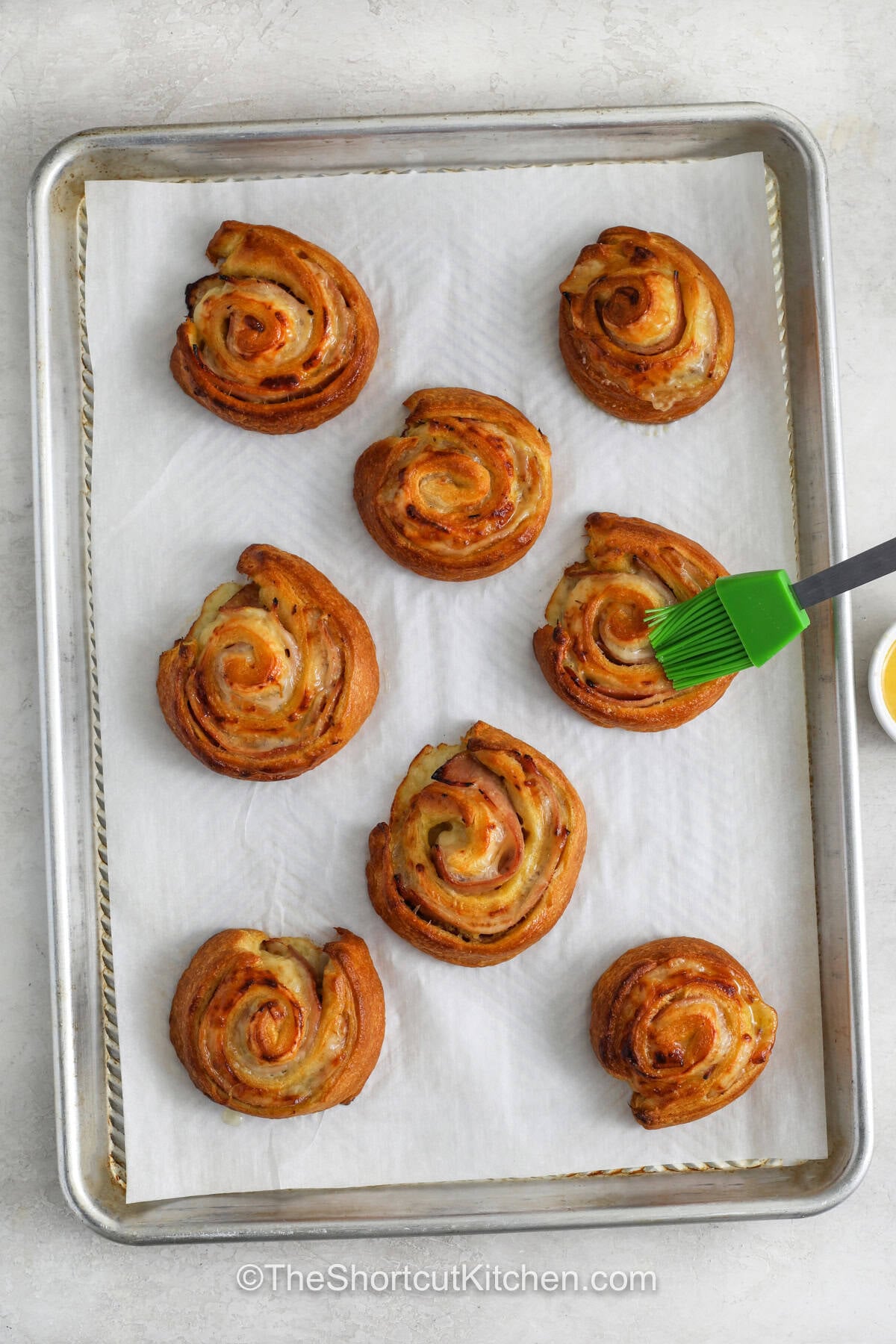 Ham and cheese pinwheels turn a few simple ingredients into a delicious snack or appetizer. Crescent roll dough is layered with honey mustard, ham, and cheese, then rolled into easy-to-serve pinwheels. After baking, a brush of garlic butter adds savory flavor to the golden tops. These tasty bites are great for parties, potlucks, brunch, or after-school snacks. Make these easy pinwheels for your next gathering. #theshortcutkitchen Ham and Cheese Pinwheels on a sheet pan brushed with melted butter