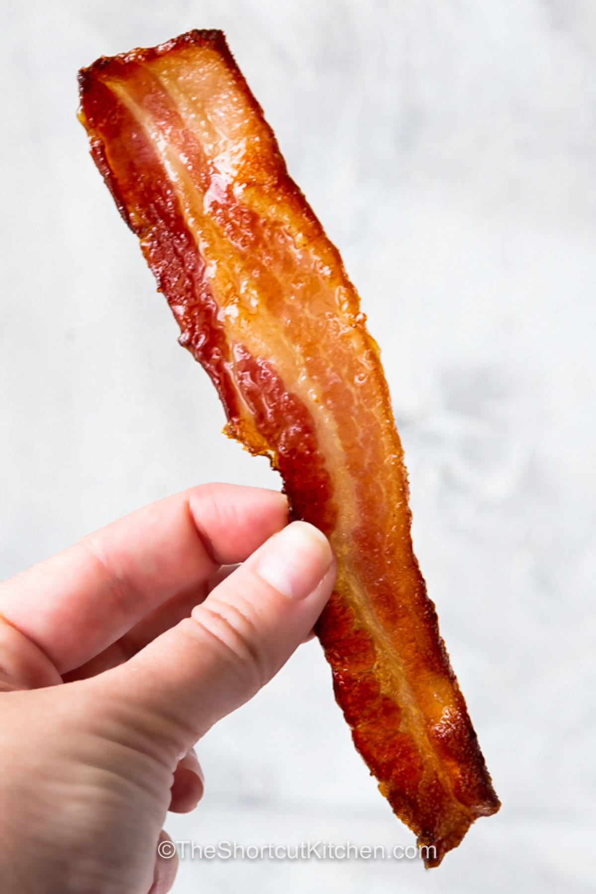 Make your next breakfast extra special with brown sugar bacon and its irresistible sweet and savory flavor. Brown sugar adds a delicious caramelized coating to crisp bacon without requiring a long list of ingredients. This easy candied bacon is perfect beside eggs, pancakes, waffles, or French toast. It also makes a tasty addition to brunch spreads and holiday breakfasts. Give it a try today. #theshortcutkitchen a hand holding a piece of crispy brown sugar bacon.
