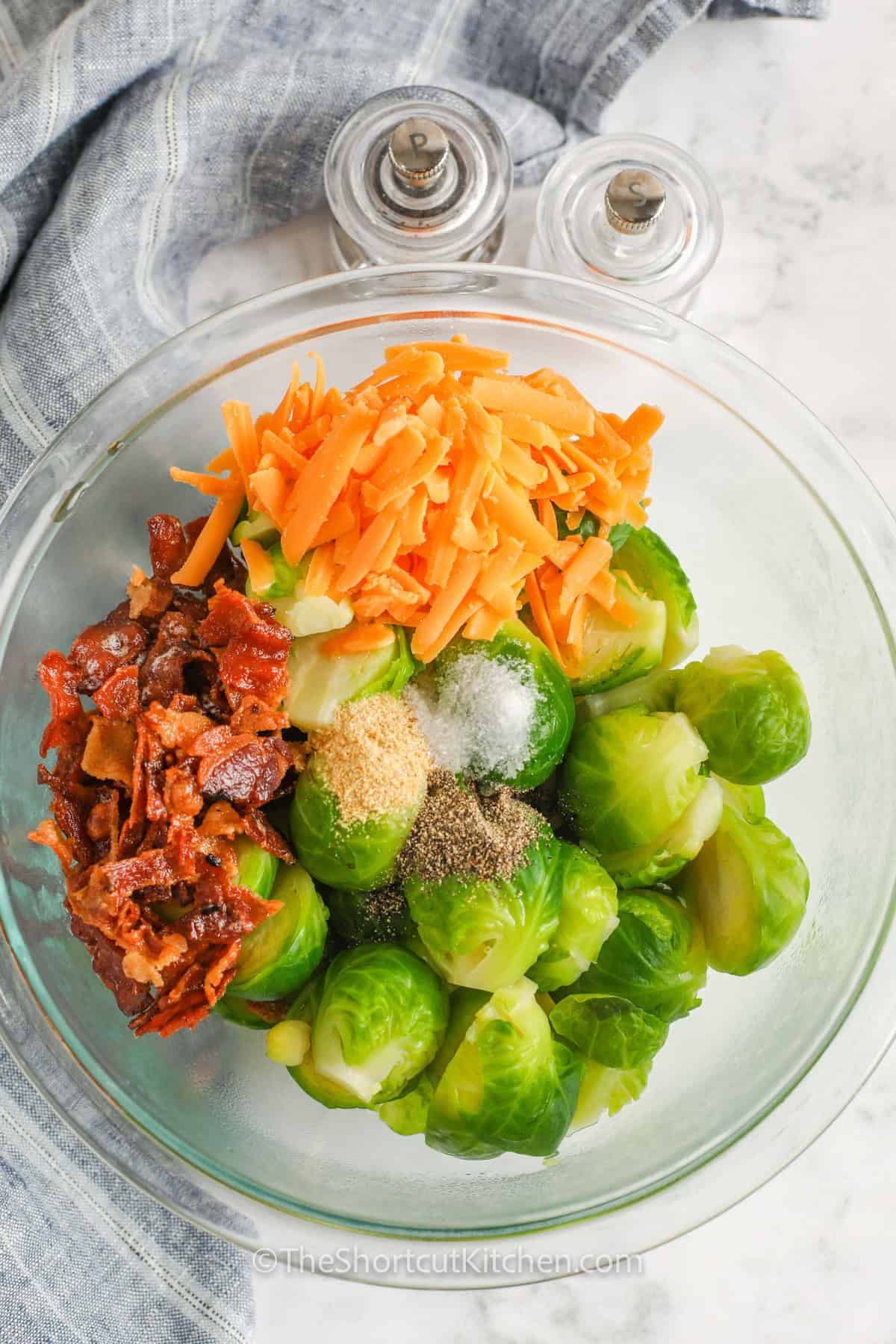 This Brussel sprout casserole with bacon has everything a great side dish needs, including tender vegetables, creamy cheddar, smoky bacon, and golden panko crunch. Fresh Brussels sprouts are precooked for faster baking, making this cheesy Brussels sprout casserole an easy 30-minute recipe. It is perfect beside chicken, pork, ham, or turkey. Pin this quick vegetable side for an easy dinner idea. #theshortcutkitchen adding seasoning to bacon , cheese and Brussels sprouts to make Brussel Sprout Casserole.
